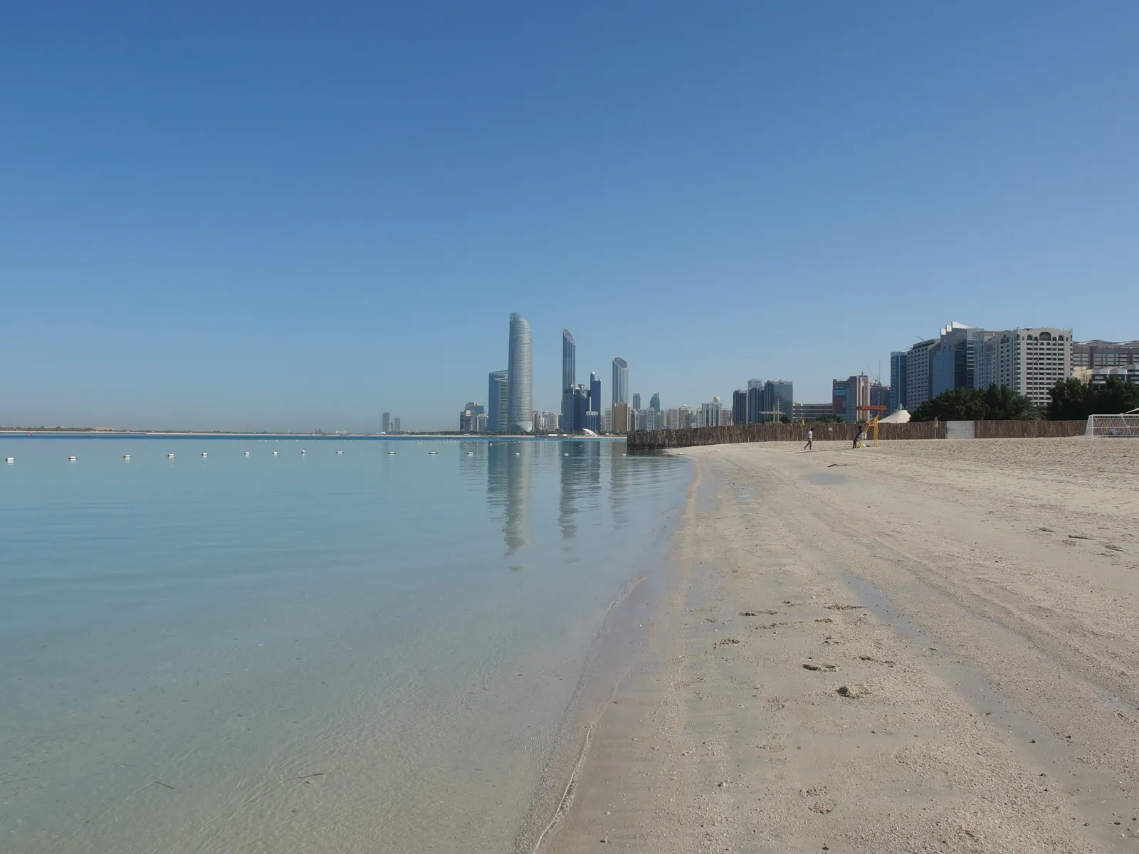 The white sand and shallow green water of Corniche Beach in Abu Dhabi, with the towers of the city standing along the shoreline in the distance