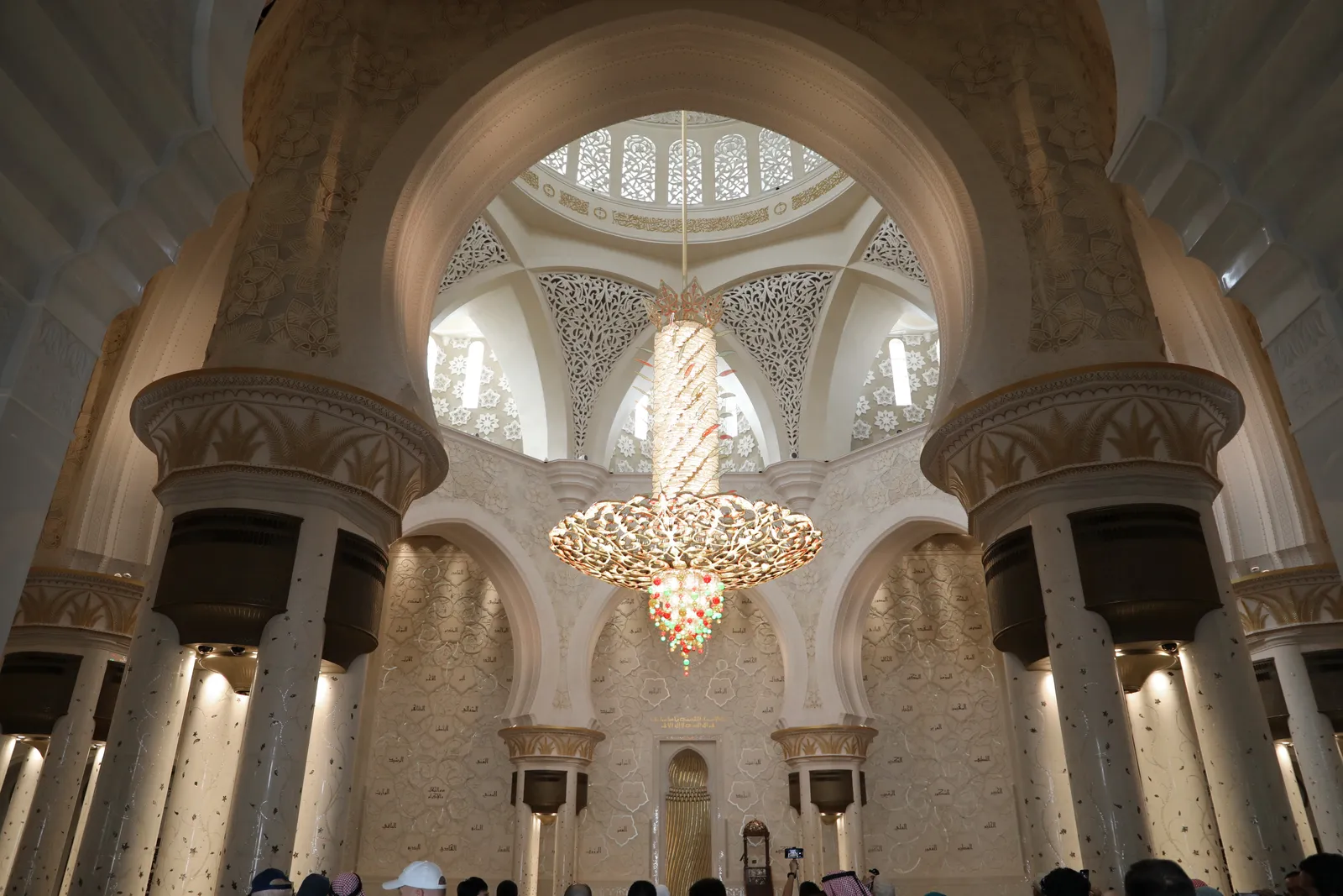 The main prayer hall of Sheikh Zayed Grand Mosque, a gold and crystal chandelier hanging under the central dome above pale marble arches and inlaid columns