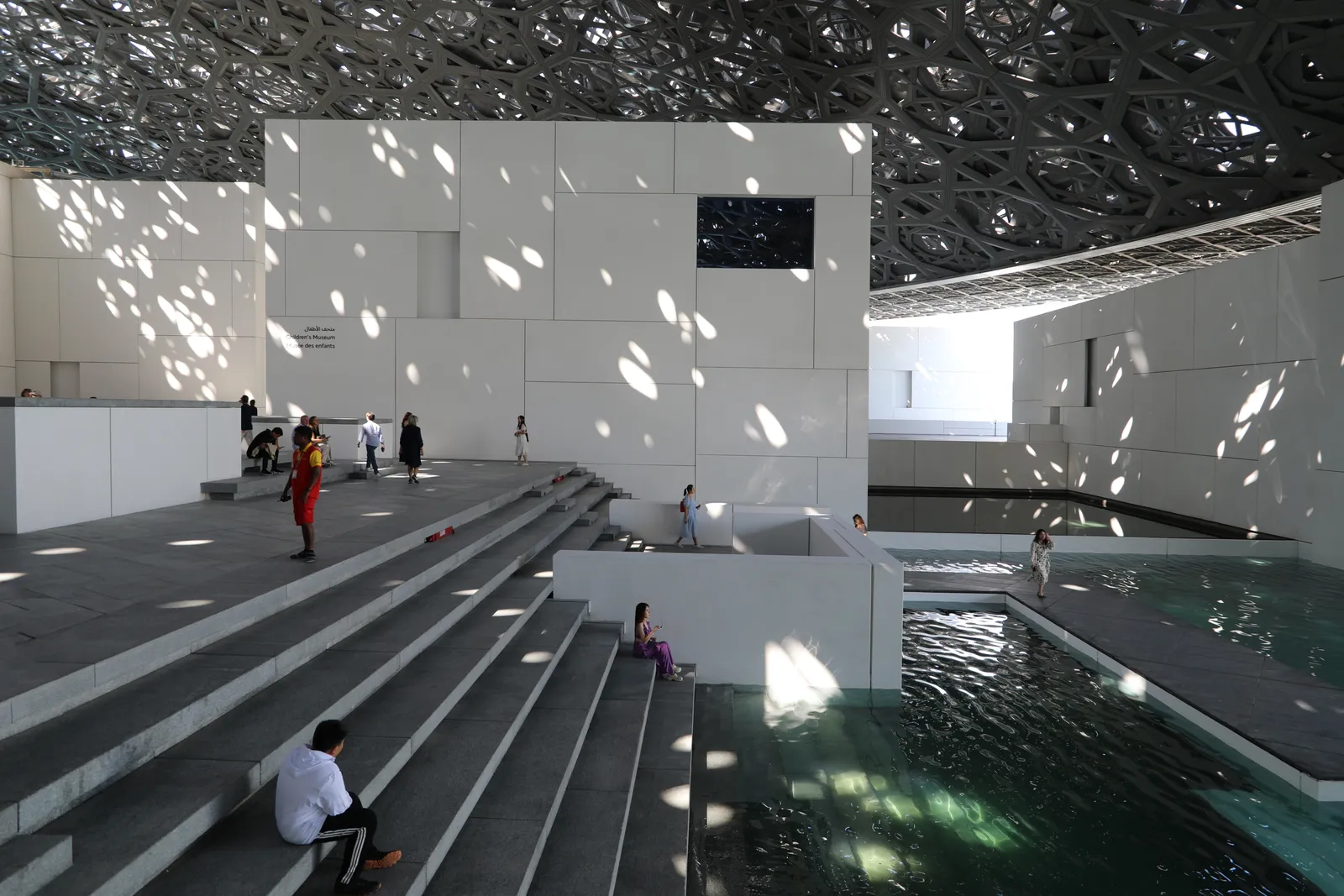 Inside Louvre Abu Dhabi, the perforated steel dome overhead scattering points of sunlight across white cubic buildings, grey steps and a channel of seawater