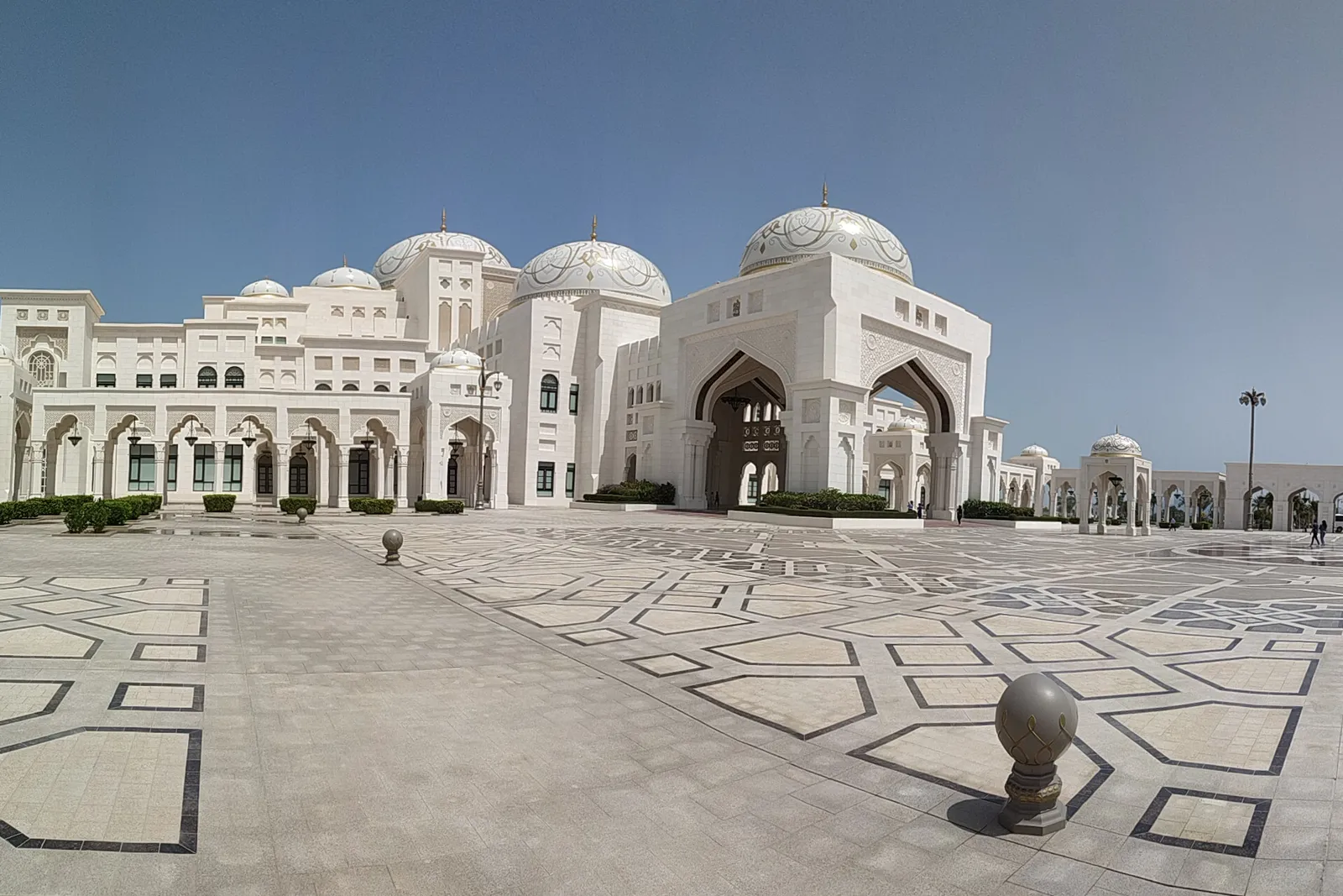 The white domes and tall pointed-arch gateway of Qasr Al Watan in Abu Dhabi, seen across a forecourt paved in a grey and white geometric pattern