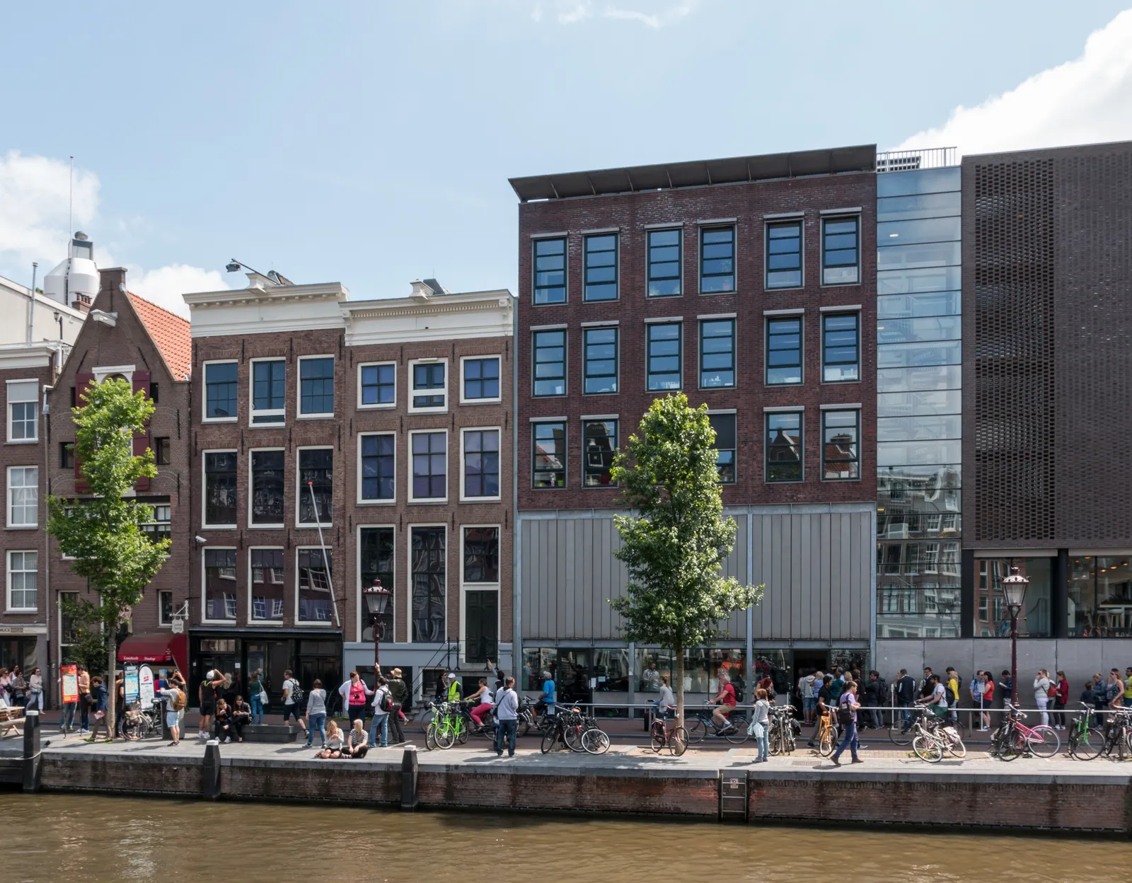 The Anne Frank House on Prinsengracht in Amsterdam, a row of canal houses beside the water with crowds and cyclists gathered along the quay outside its entrance