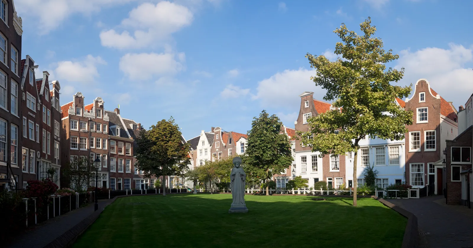 The enclosed Begijnhof courtyard in Amsterdam, a wide green lawn ringed by low historic houses with stepped and bell gables, with a stone statue of a beguine in the middle