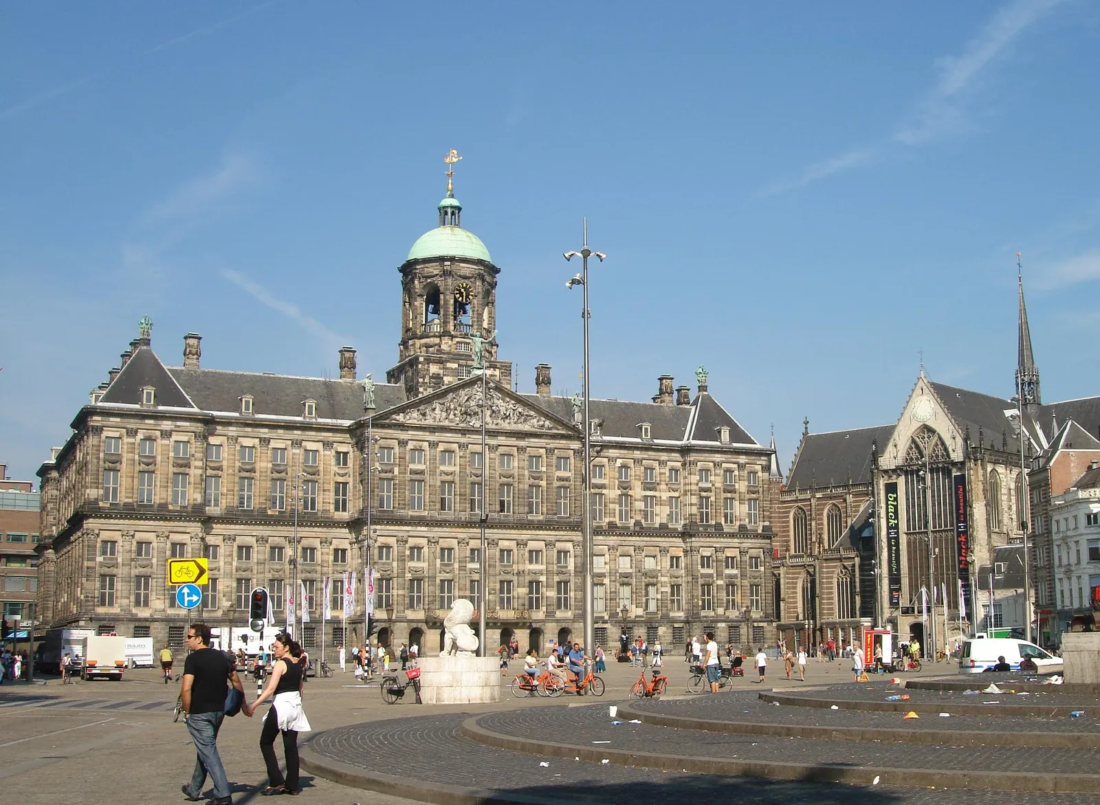 The Royal Palace and the Nieuwe Kerk seen across the open paving of Dam Square in Amsterdam on a clear summer day, with cyclists and pedestrians crossing in front