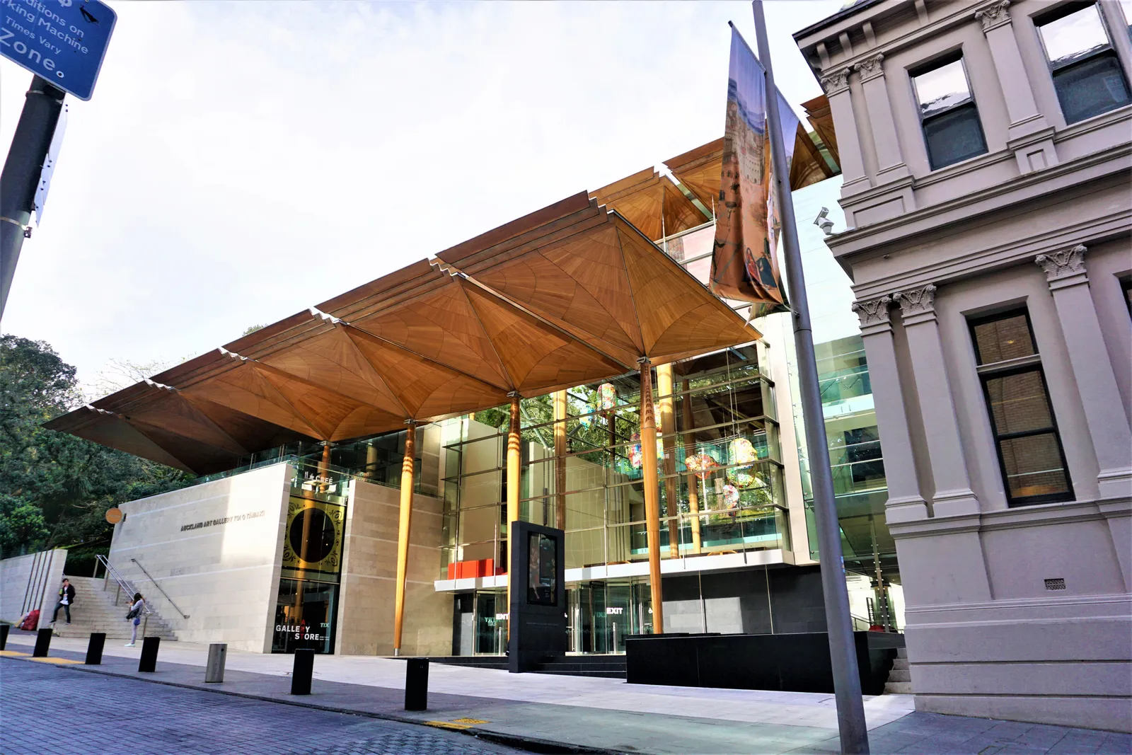 The entrance to Auckland Art Gallery Toi o Tāmaki, a canopy of tall timber columns spreading into a roof over the forecourt, with the older stone wing of the gallery beside it