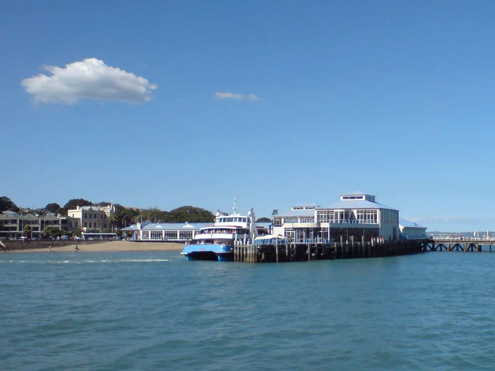 Devonport wharf across the water with a passenger ferry moored alongside, the white ferry building at the end of the pier and a beach and trees behind it
