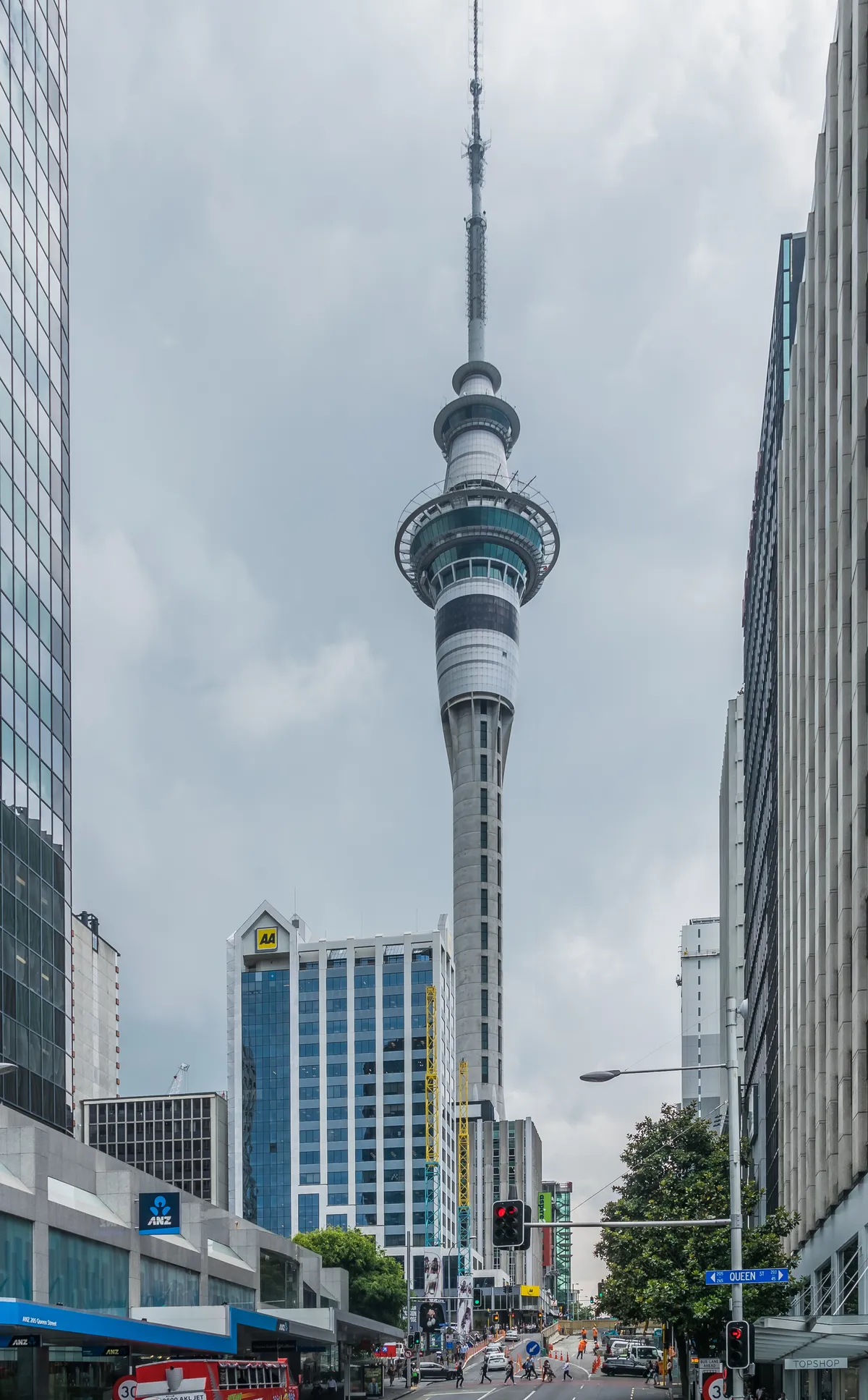 The Sky Tower seen down a central Auckland street, its observation pods and slender mast rising above the office buildings on either side with traffic and pedestrians at the crossing below