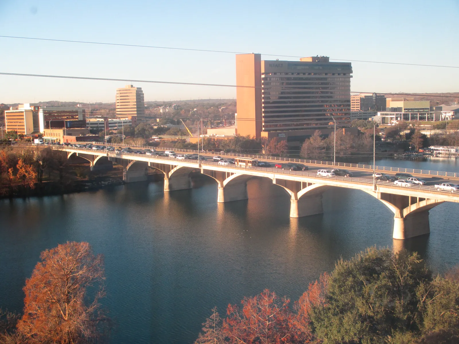 The Congress Avenue bridge in Austin seen from upstream, a row of concrete arches carrying four lanes of traffic across Lady Bird Lake, with office buildings on the far bank and autumn trees in the foreground