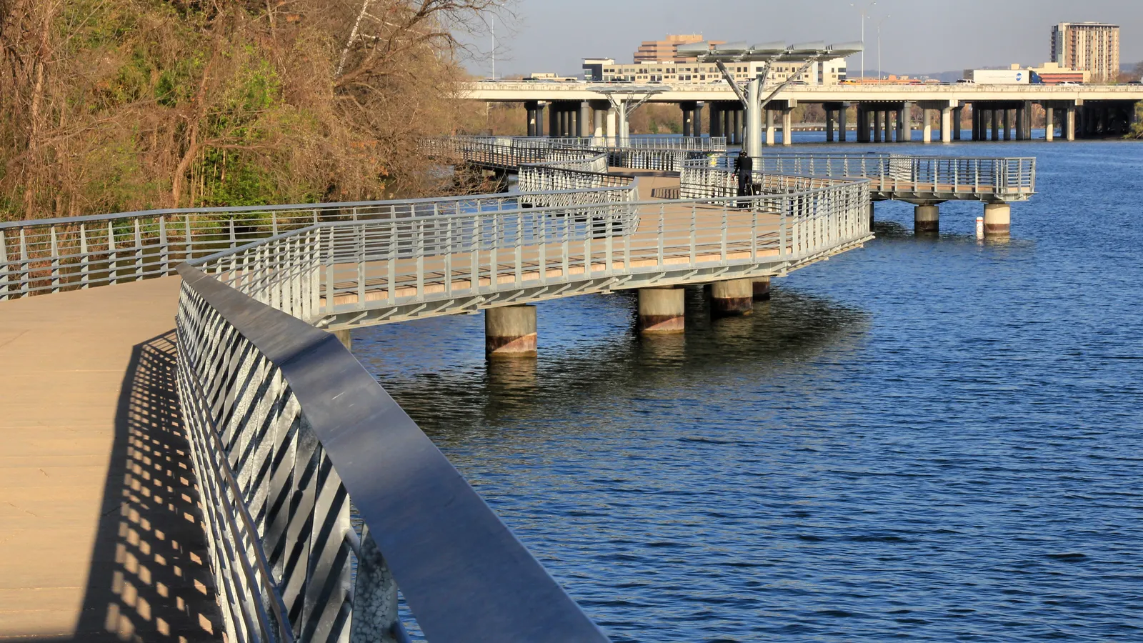 The Boardwalk on Lady Bird Lake in Austin, a wide concrete walkway on piers curving out over the water behind a steel railing, with a road bridge and low buildings in the distance