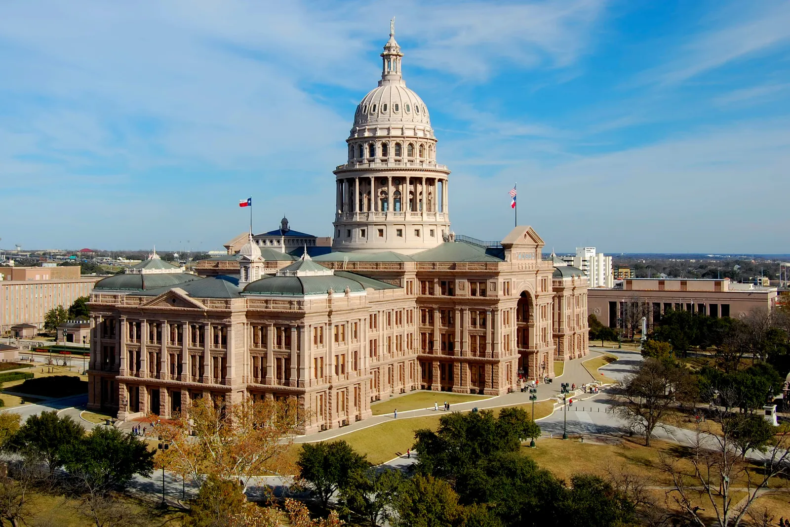 The Texas State Capitol in Austin seen from above and to one side, a long pink granite building with a tall domed rotunda at its center and a statue on top, standing in lawns and trees