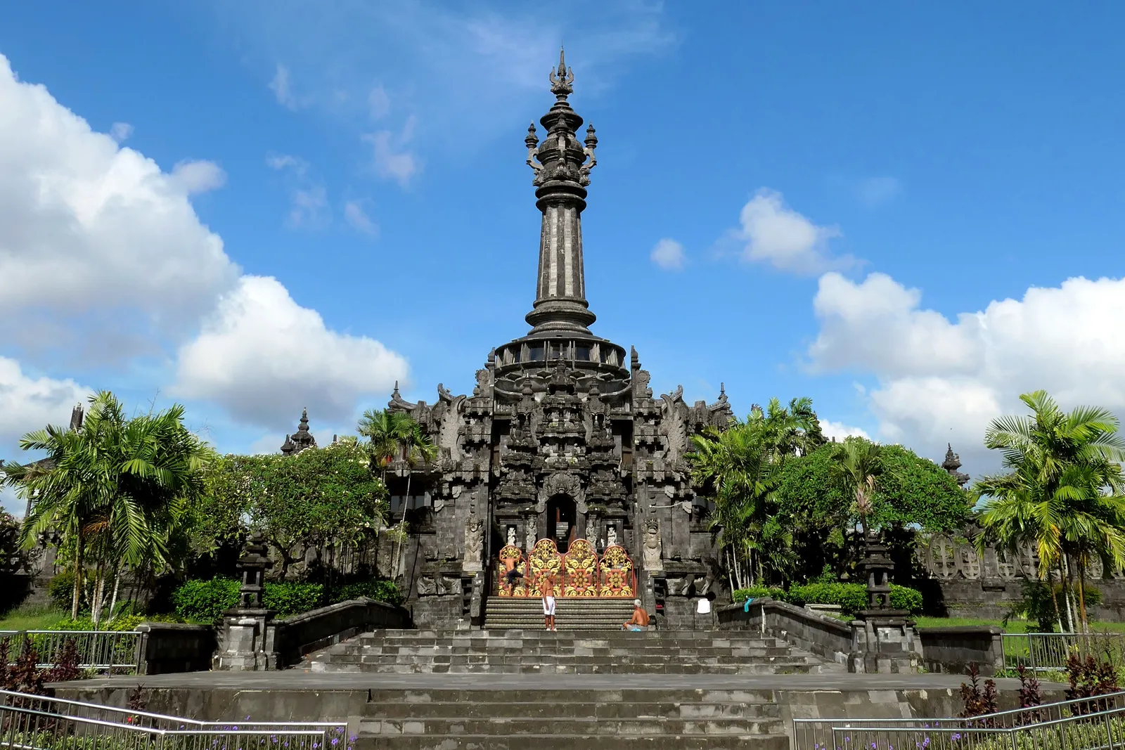 The Bajra Sandhi Monument in Denpasar, a dark stone monument with a tall fluted spire rising from a broad carved base, with palms and steps in front of it
