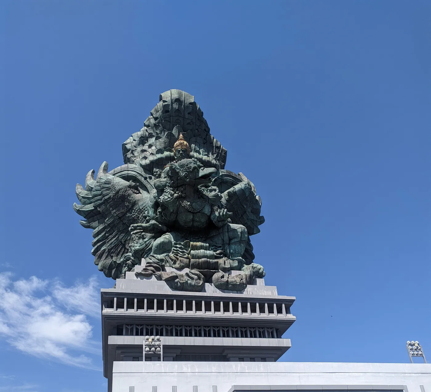 The green copper Garuda Wisnu Kencana statue, Vishnu riding the eagle Garuda, seen from below on its tiered white pedestal against a clear blue sky