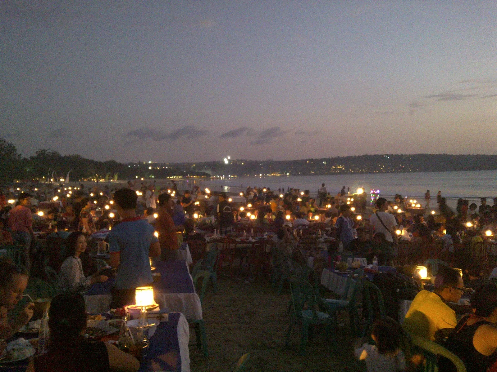 Hundreds of candlelit dinner tables set out on the sand along Jimbaran Bay at dusk, with the lights of the bay curving away behind them