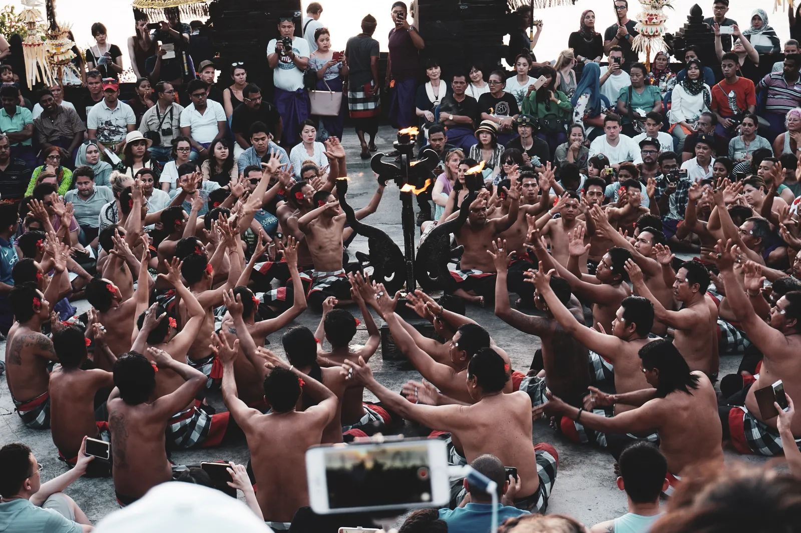 A ring of bare-chested Kecak chanters sitting with their arms raised around a lit candelabra, surrounded by rows of seated spectators in the open-air amphitheatre at Uluwatu