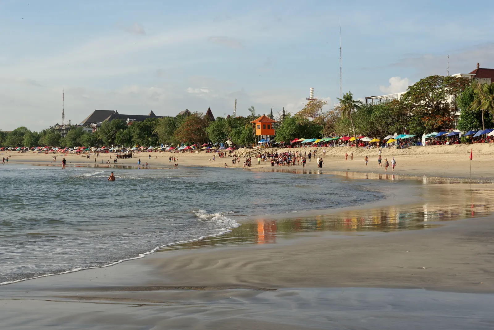 Wet sand and shallow surf along Kuta Beach in Bali, with crowds, coloured umbrellas and palm trees lining the shore in the late afternoon