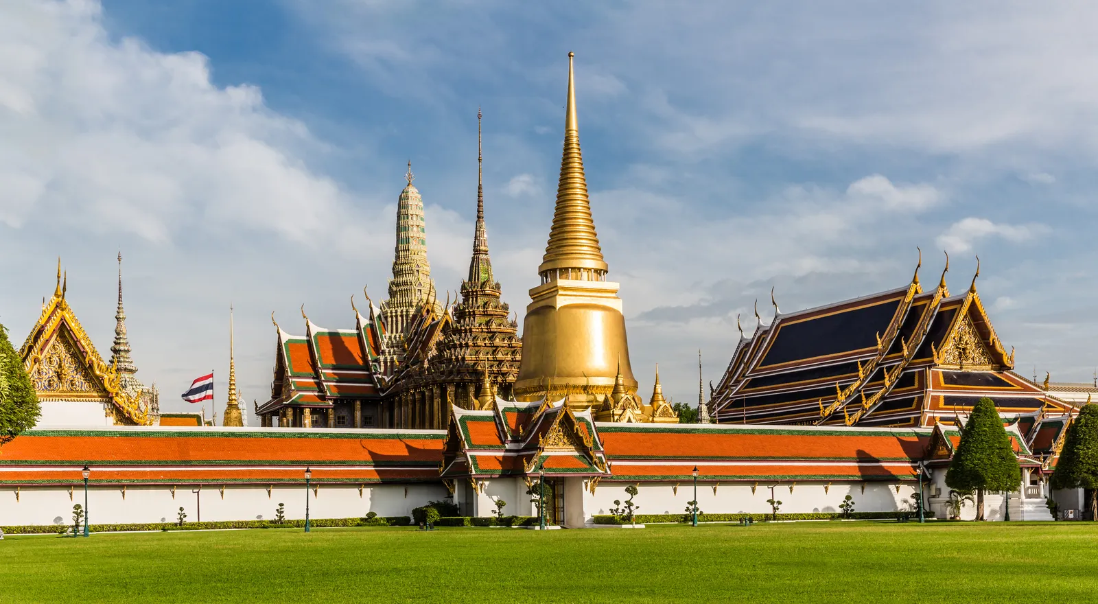 Wat Phra Kaew inside the Grand Palace in Bangkok, a golden bell-shaped chedi flanked by tiered orange and green temple roofs behind a long white wall, seen across a mown lawn