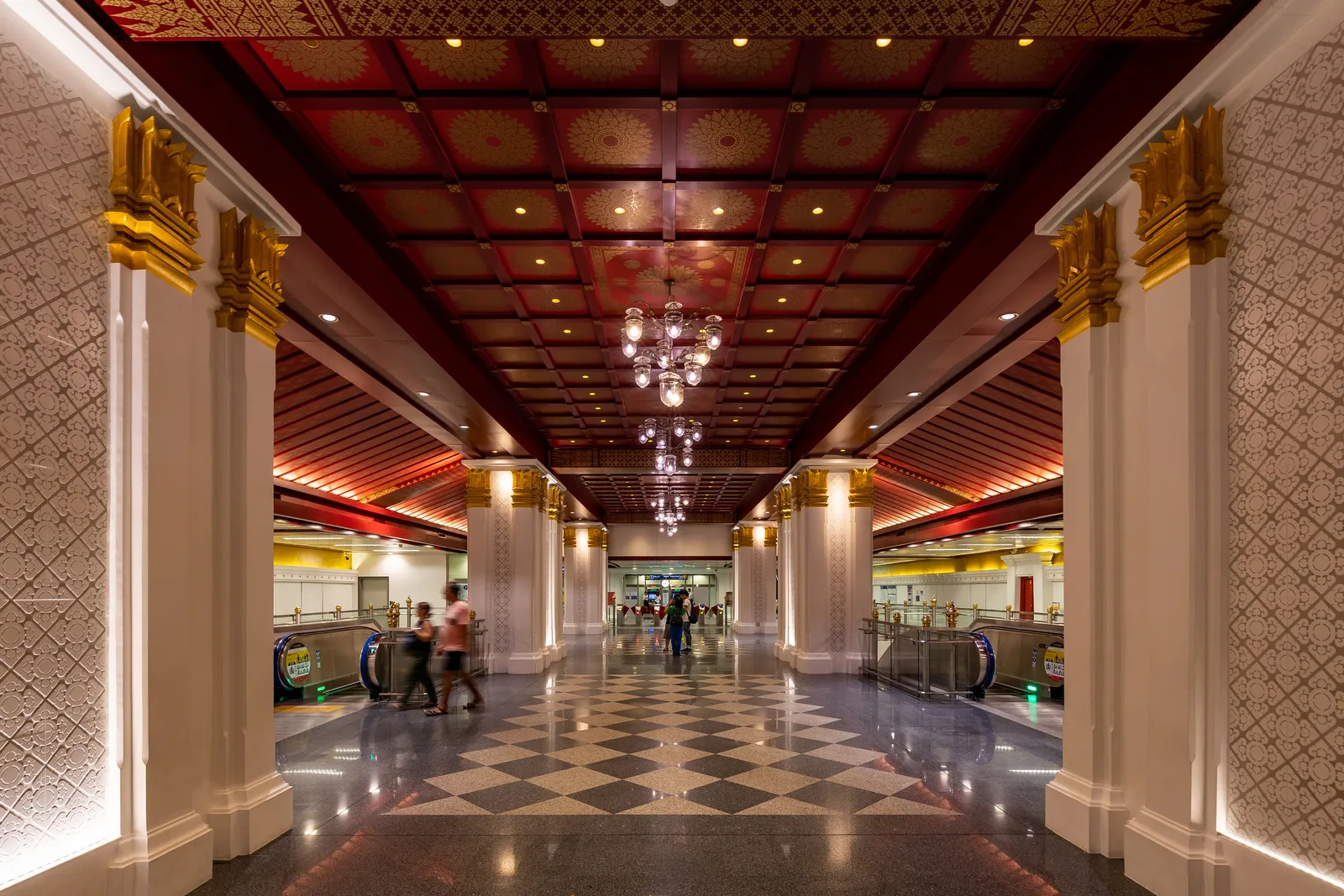 The concourse of Sanam Chai metro station in Bangkok, white columns with gilded capitals under a red and gold coffered ceiling hung with chandeliers, escalators on both sides and a black and white tiled floor