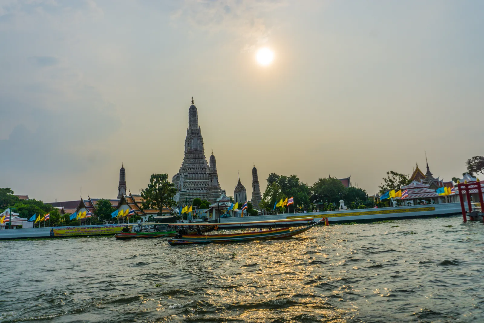 Wat Arun seen from a boat on the Chao Phraya in Bangkok, its tall pale tapering tower and four smaller towers rising above the trees on the far bank with long-tail boats crossing in front
