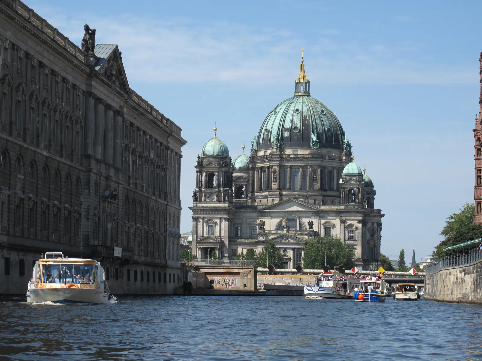 The green copper dome of the Berliner Dom rising above Museum Island in Berlin, seen from a boat on the Spree with the colonnaded flank of the island on the left and tour boats on the water