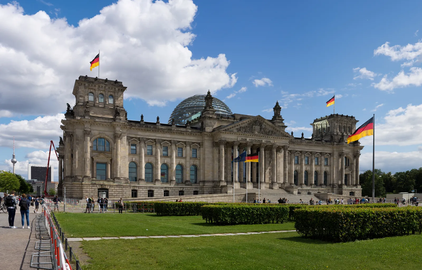The west front of the Reichstag building in Berlin with German and European flags flying, Norman Foster's glass dome visible above the roofline, and the lawn of the Platz der Republik in the foreground