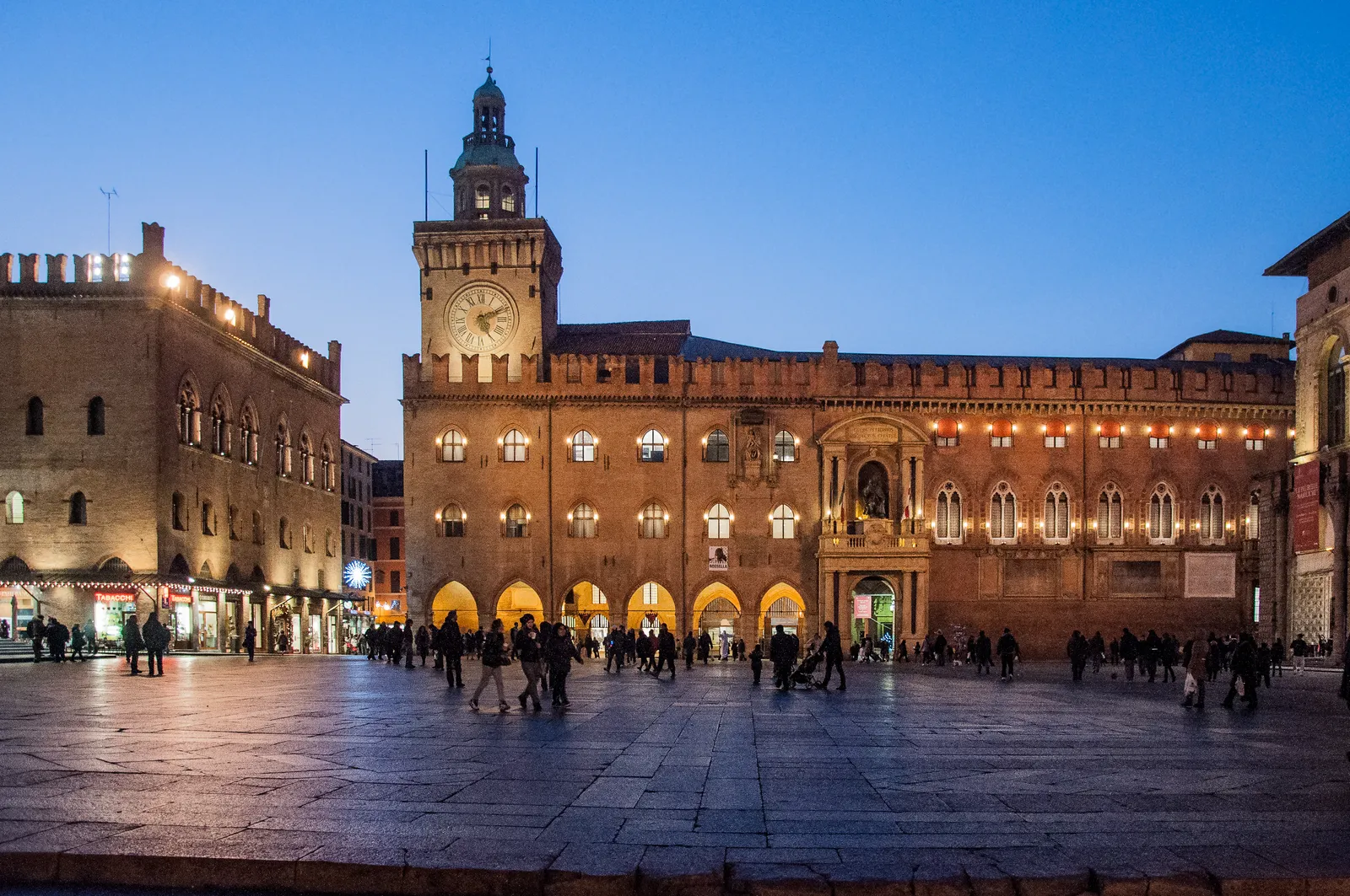 Palazzo d'Accursio, Bologna's city hall, lit up at dusk on Piazza Maggiore, its brick clock tower rising above a long crenellated facade