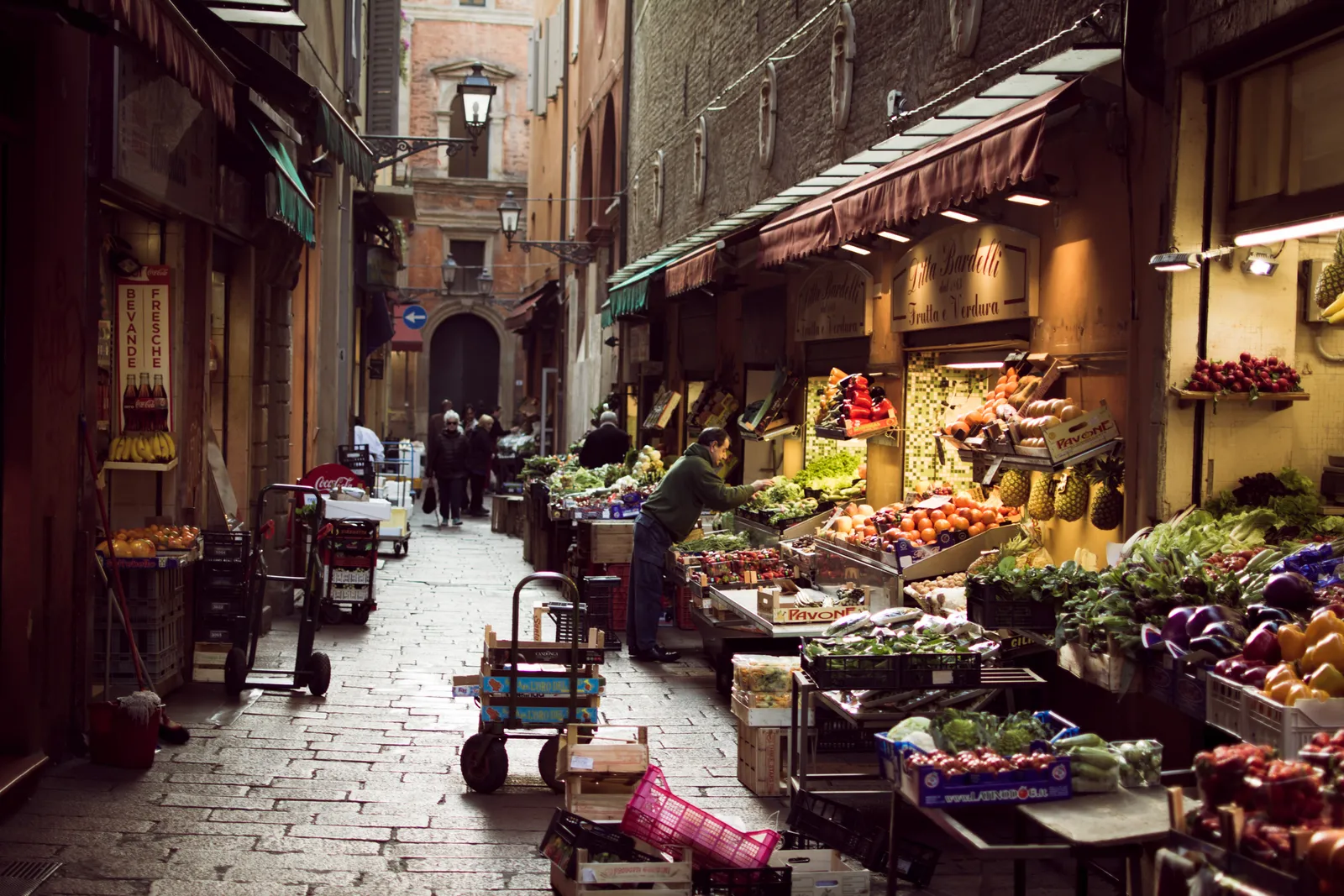 Crates of fruit and vegetables set out under shop awnings along Via Pescherie Vecchie, a narrow stone-paved lane in Bologna's Quadrilatero market district