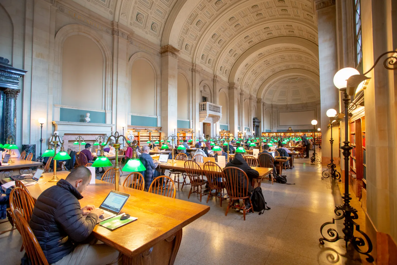 Bates Hall at the Boston Public Library, a long barrel-vaulted reading room lined with oak tables and green-shaded lamps, with readers working under the arched ceiling