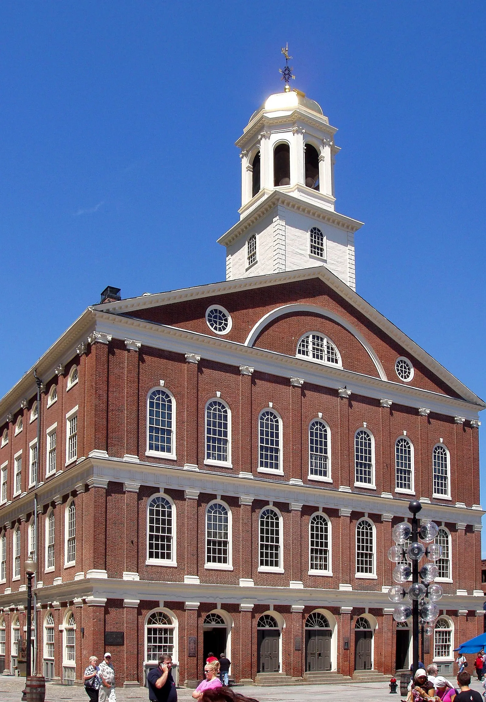 Faneuil Hall's brick Georgian facade in Boston, with a white cupola and gilded grasshopper weathervane rising above the gabled roof and pedestrians gathered on the plaza in front