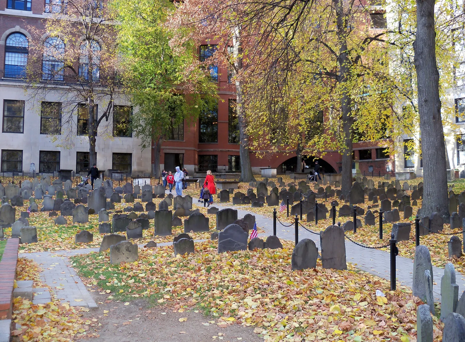 Rows of thin slate headstones in Boston's Granary Burying Ground under autumn leaves, with the brick and stone buildings of Tremont Street behind