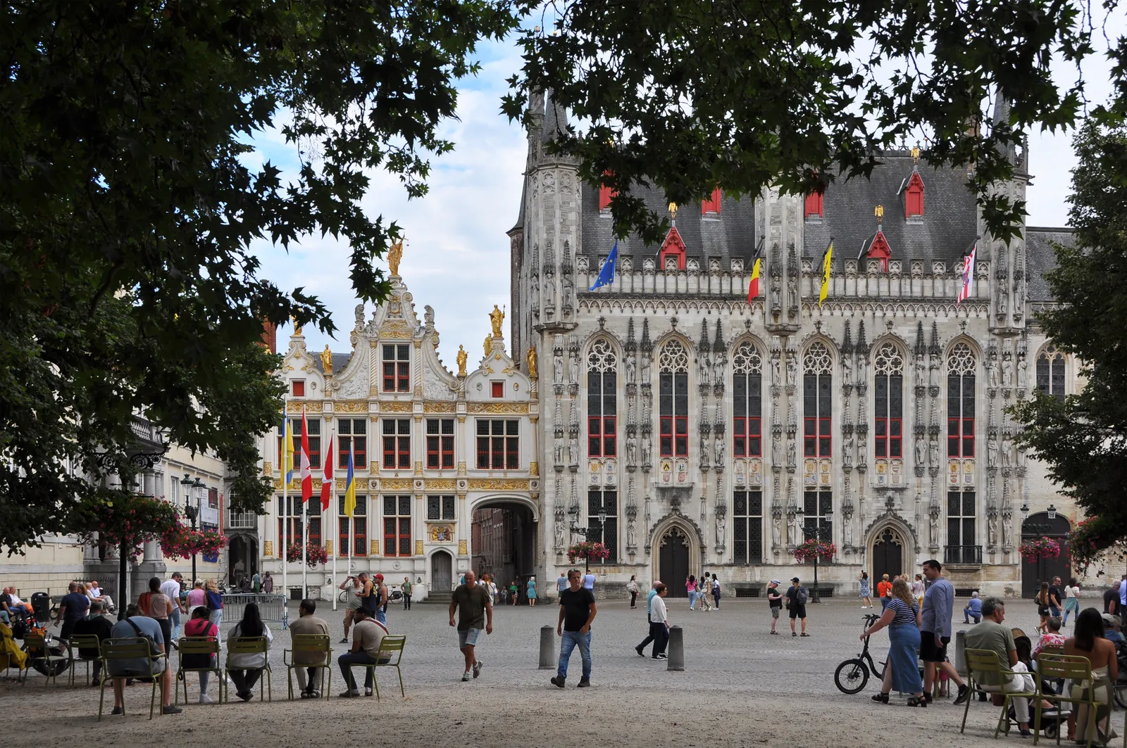 Burg square in Bruges, the white Gothic city hall with its turrets and statue niches beside the gilded Renaissance front of the old recorder's house, people sitting out on the open square