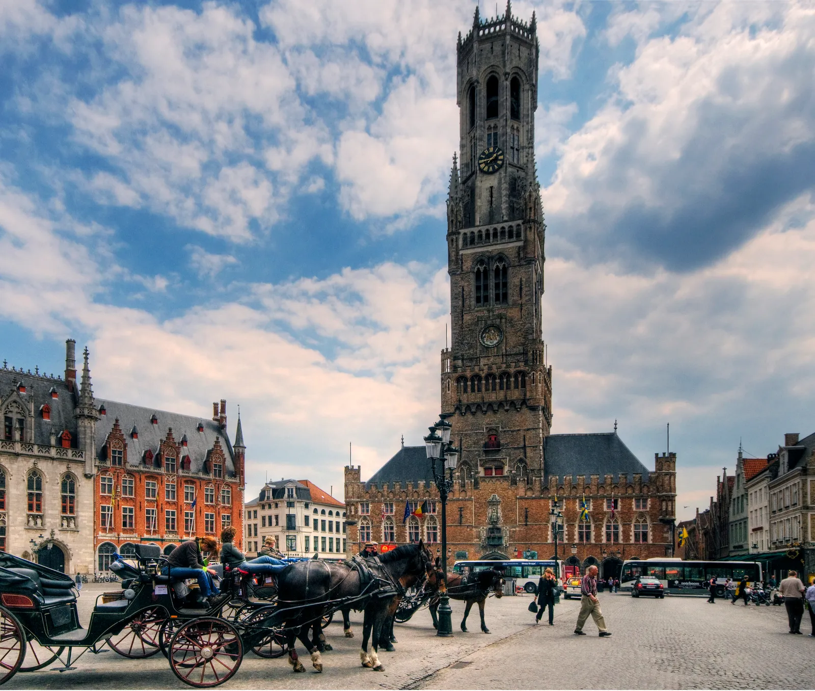 The Markt in Bruges, the brick belfry tower rising above the covered market hall with horse-drawn carriages waiting on the cobbles and a row of stepped-gable guild houses along one side