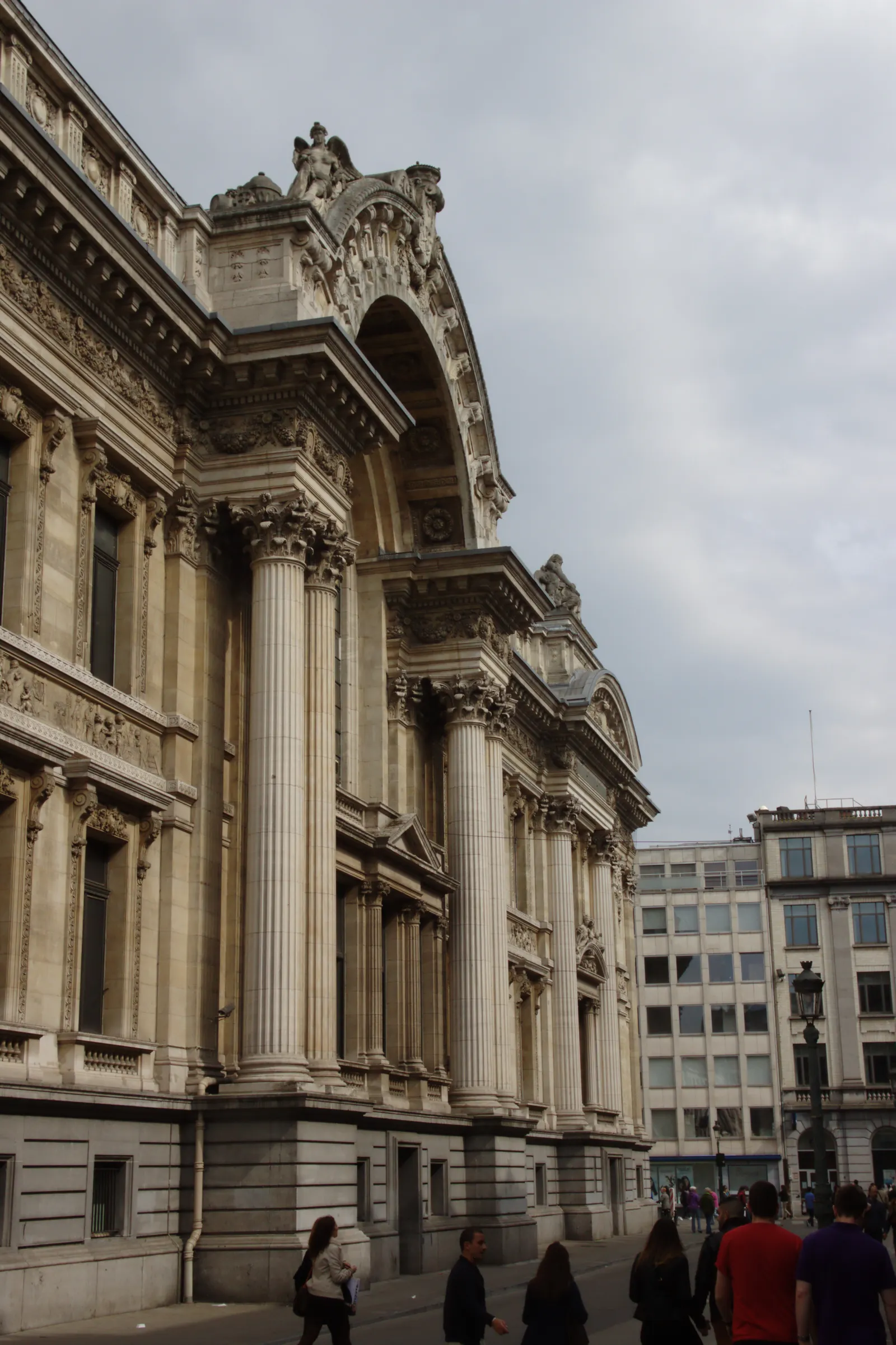 The columned front of the old Brussels Stock Exchange, its carved pediment and sculpted friezes above a row of Corinthian columns, with people walking the pedestrian street below