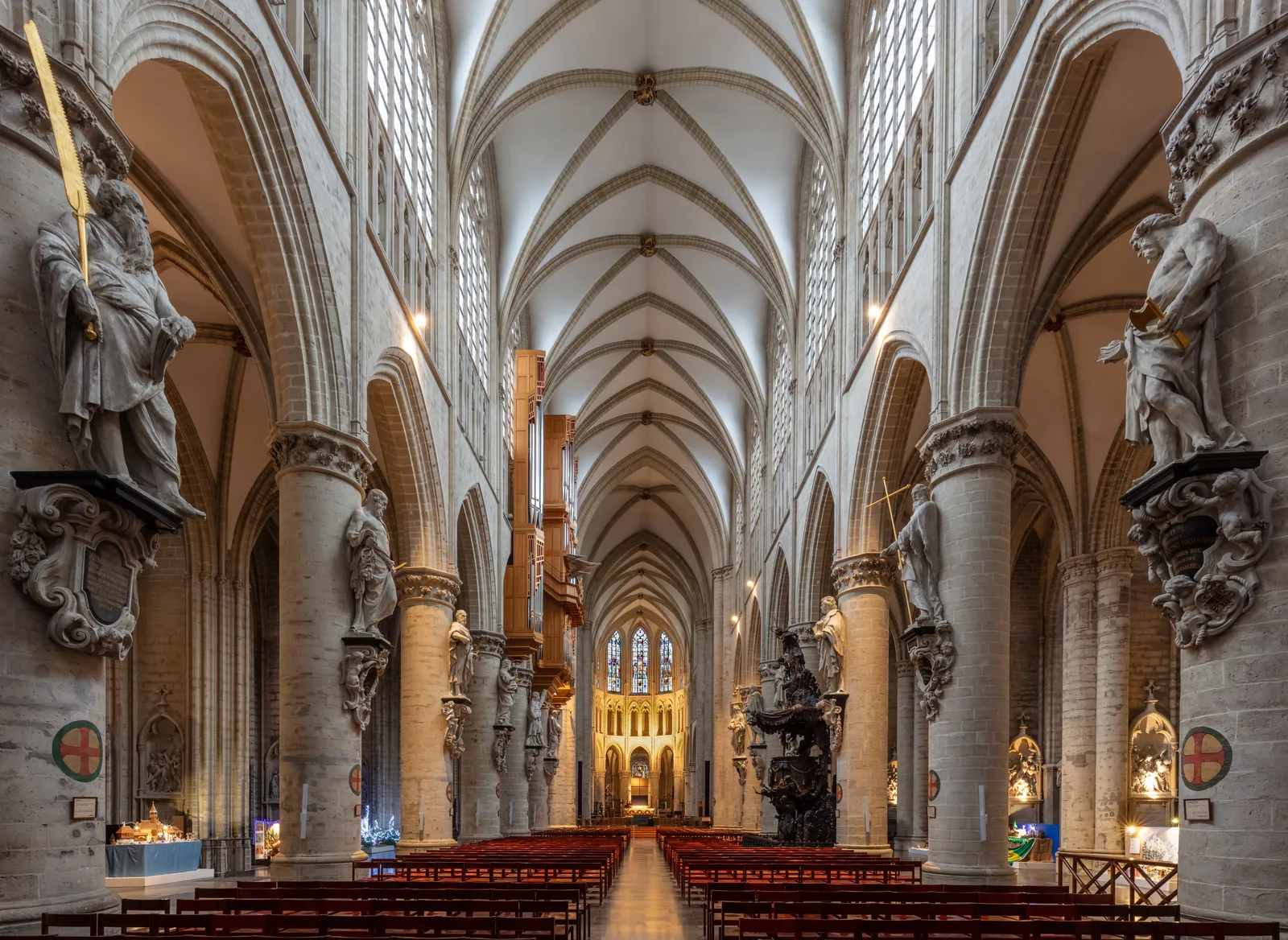 The nave of the Cathedral of St. Michael and St. Gudula in Brussels, pale stone columns carrying a ribbed Gothic vault, with carved apostle statues mounted against the columns above the pews