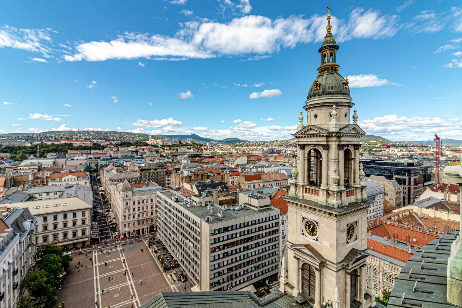 The view from the panorama terrace of St. Stephen's Basilica in Budapest, looking down past one of the basilica's stone bell towers to the square below and the rooftops of Pest, with the Buda hills on the horizon