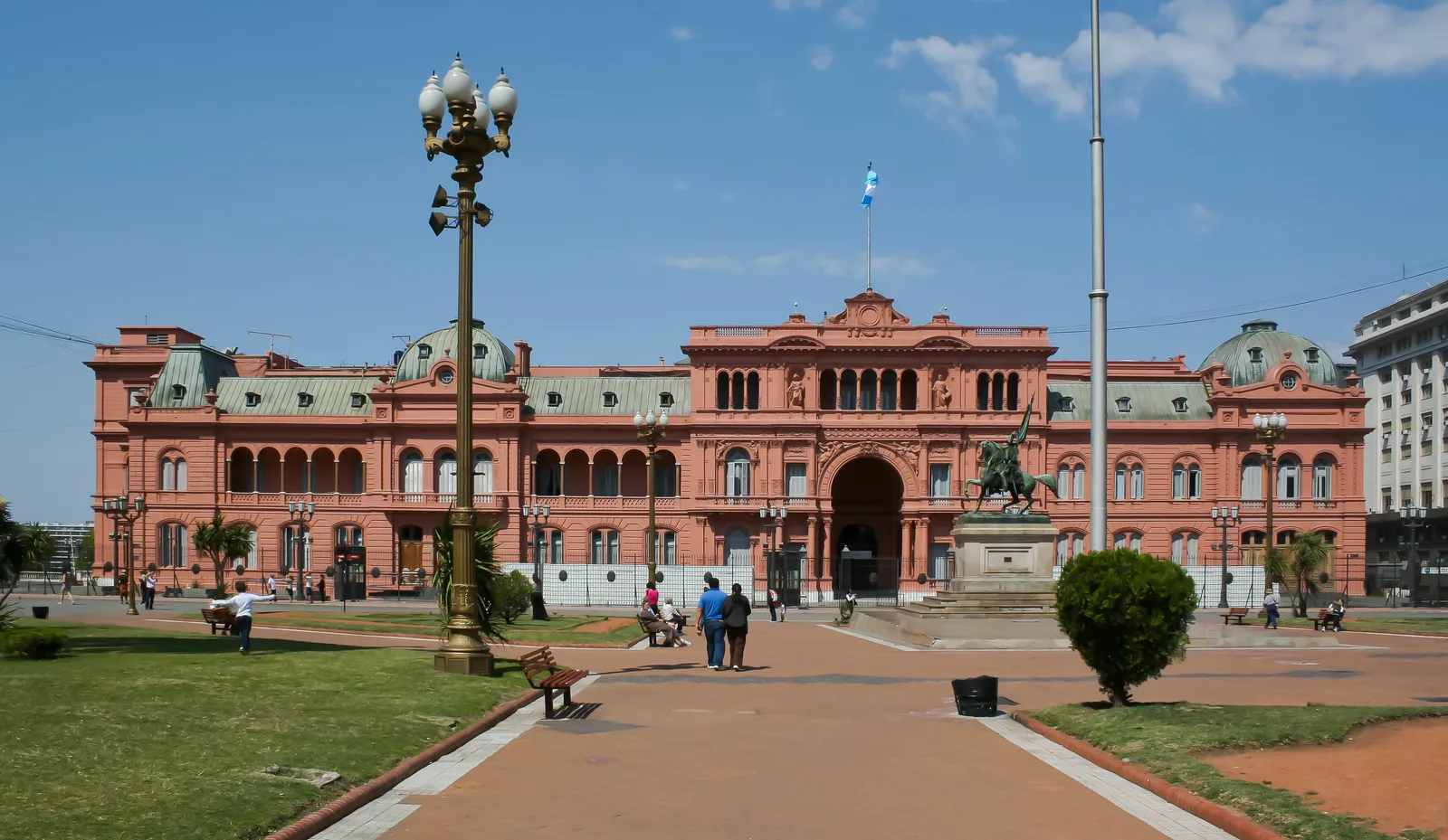 The pink facade of the Casa Rosada seen across Plaza de Mayo in Buenos Aires, with an arched central gateway, an equestrian statue in front of it and the Argentine flag flying from the roof