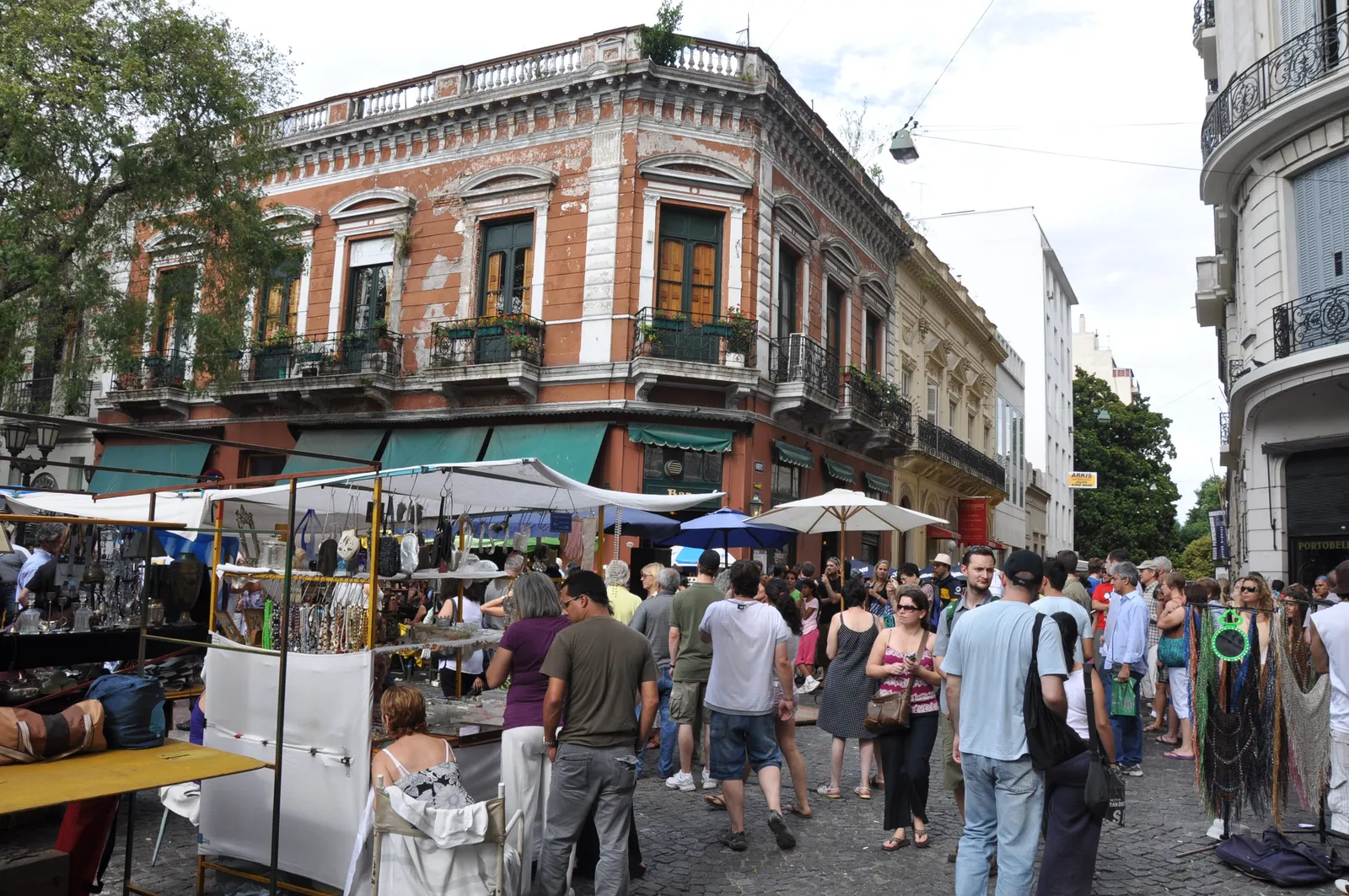 Market stalls under white umbrellas filling the cobbled street beside Plaza Dorrego in San Telmo, Buenos Aires, with crowds browsing between them and a red-brick corner building with iron balconies above