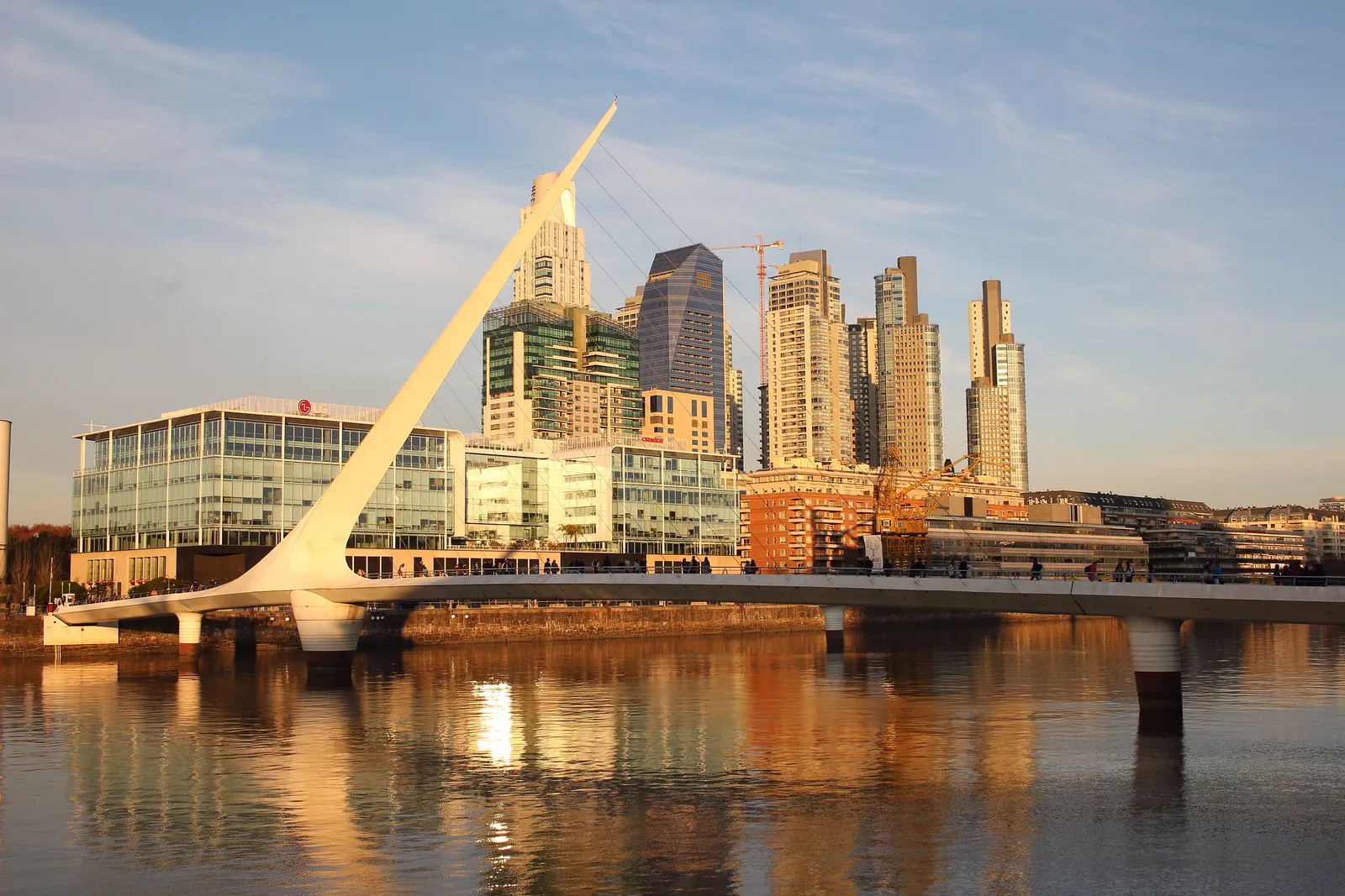 The white Puente de la Mujer footbridge across a dock in Puerto Madero, Buenos Aires, its single angled pylon leaning over the water with the district's glass towers behind it in late afternoon light