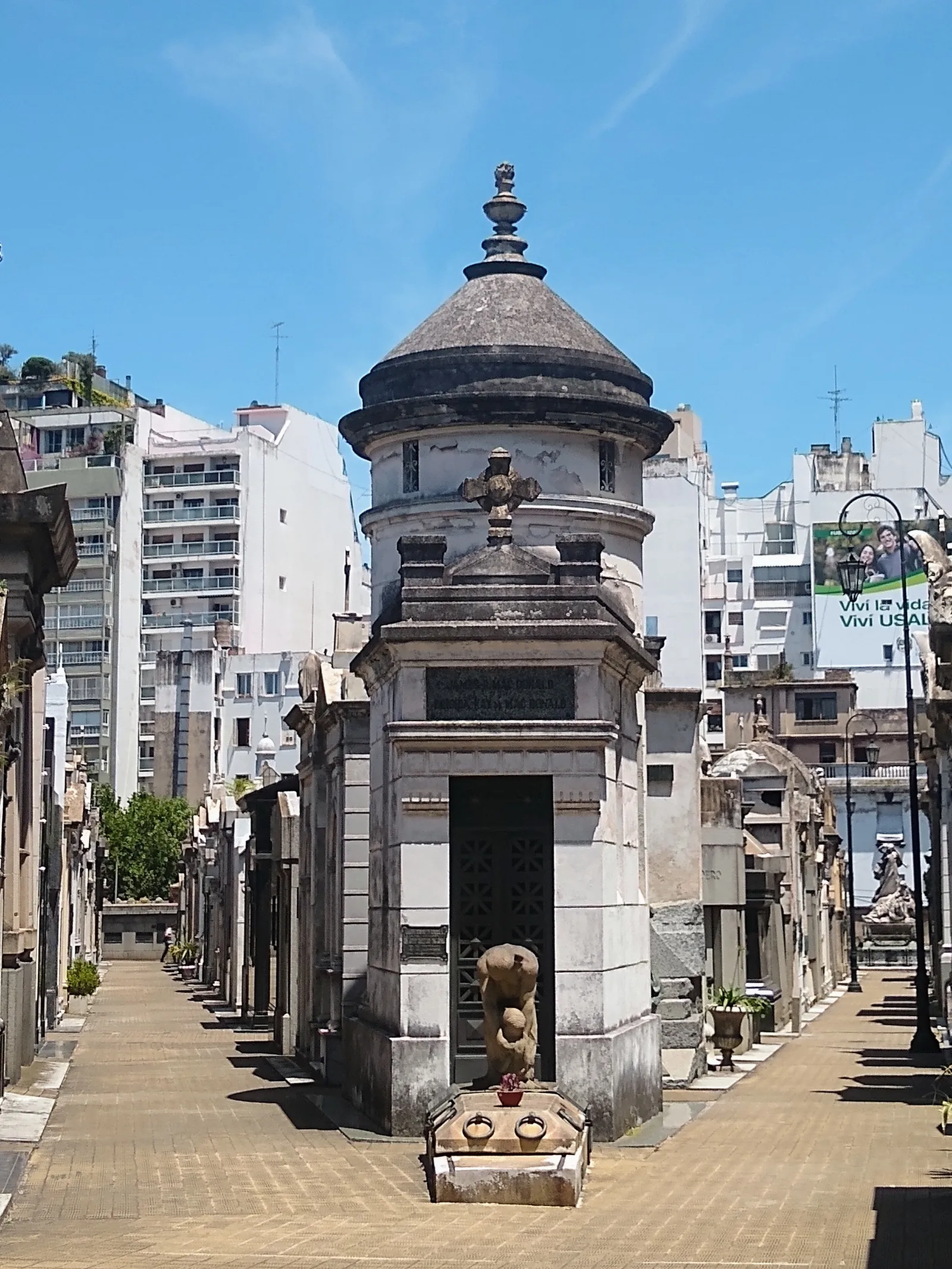A paved lane of stone mausoleums at Recoleta Cemetery in Buenos Aires, running away from the camera between rows of vaults, with the balconies of city apartment blocks rising directly behind the cemetery wall
