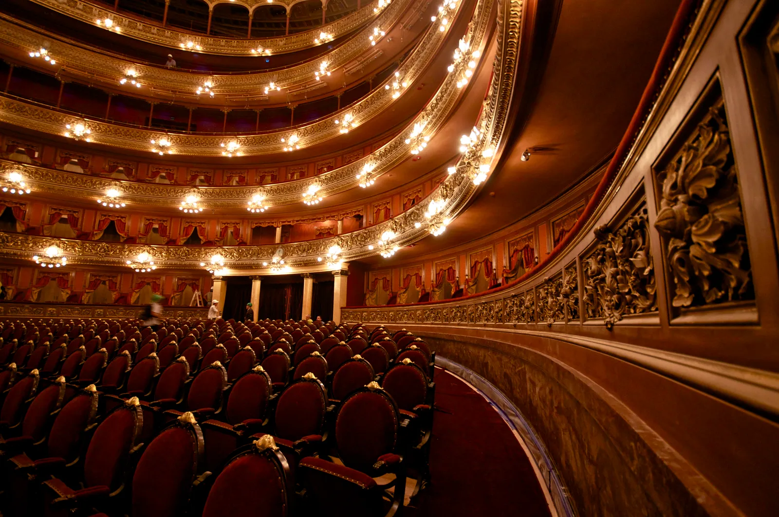 The horseshoe-shaped main auditorium of the Teatro Colón in Buenos Aires, with tiers of gilded balconies lit by rows of lamps curving around rows of empty red velvet seats