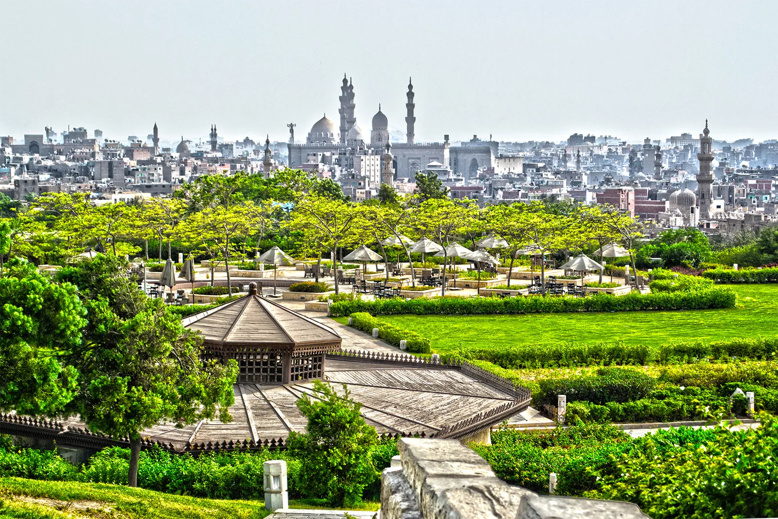 Lawns, clipped hedges and a timber pavilion in Al-Azhar Park, with the domes and minarets of a mosque complex and the flat rooftops of Cairo filling the horizon beyond