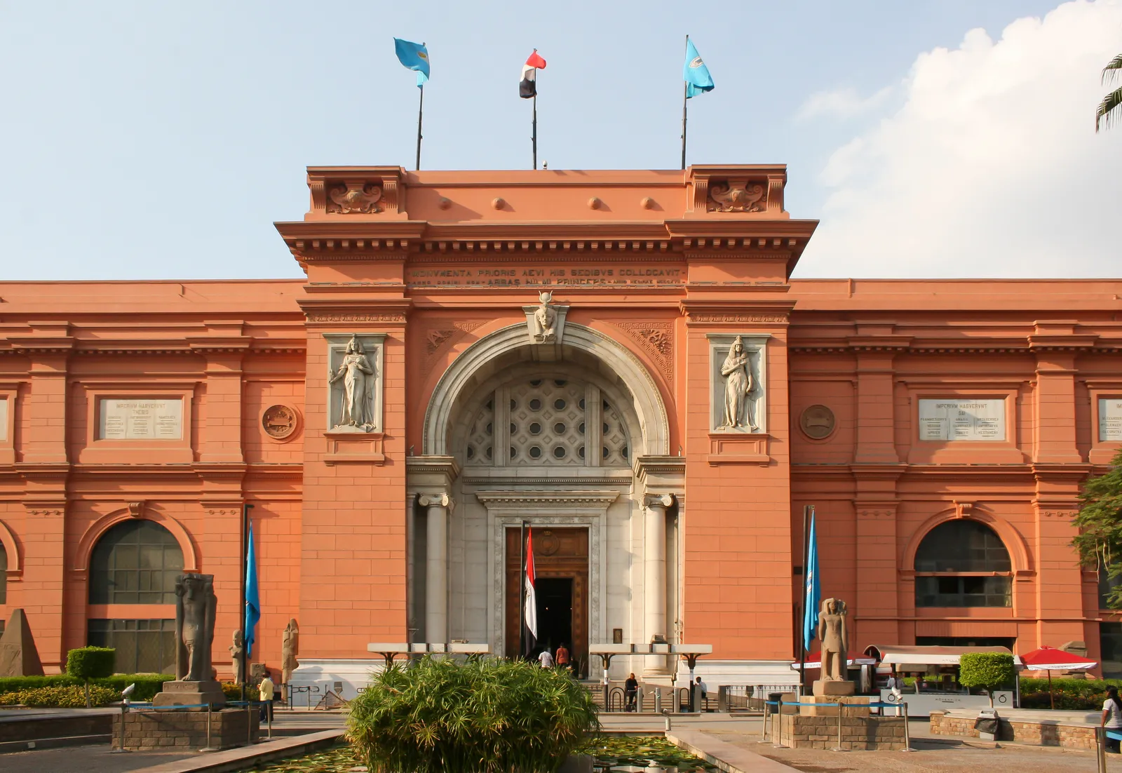 The salmon-pink neoclassical front of the Egyptian Museum on Tahrir Square, with an arched central entrance, stone figures set into niches on either side and flags flying from the roofline