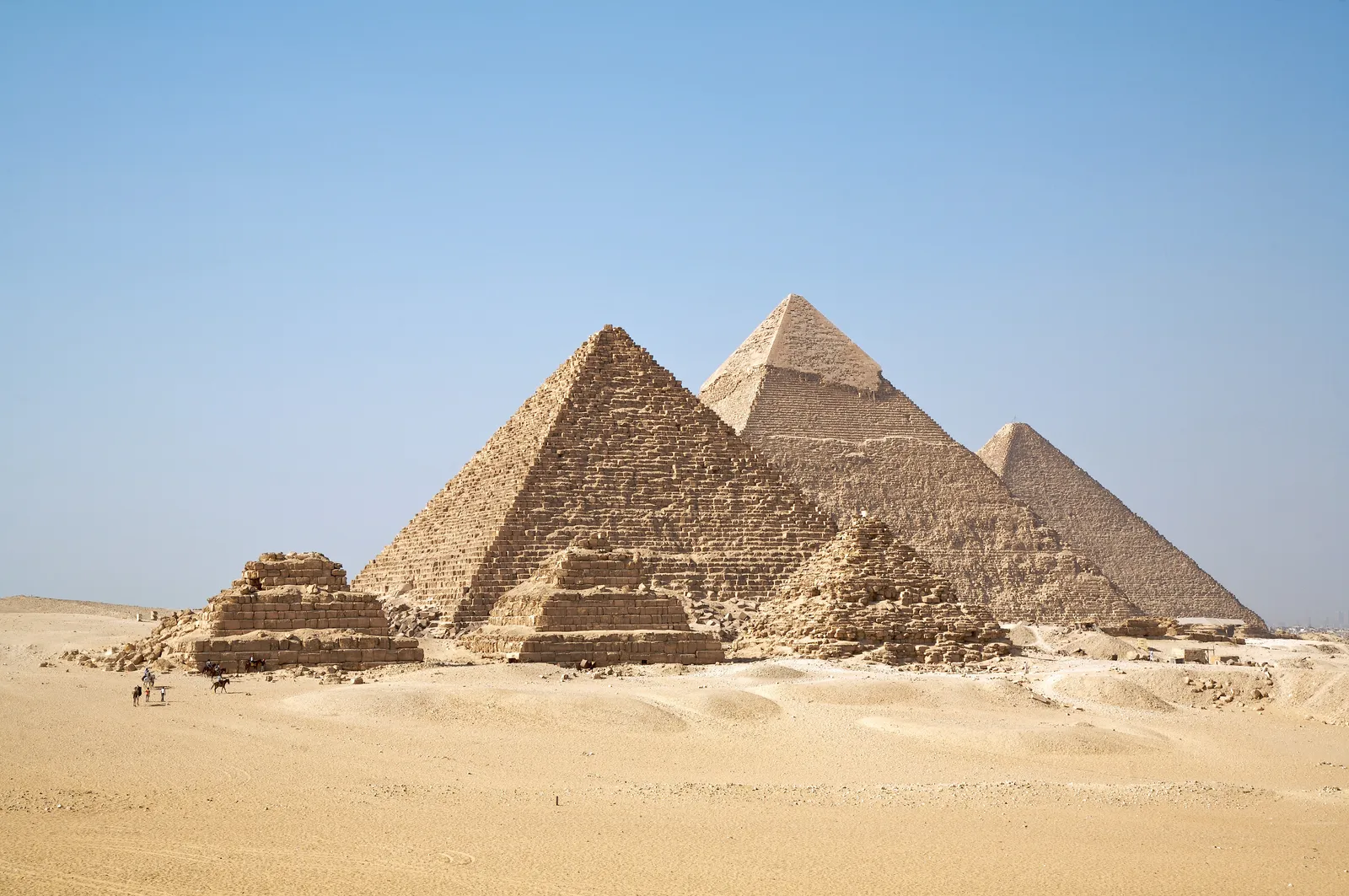 The three main pyramids of Giza lined up across the desert under a clear sky, with the smaller queens' pyramids in ruins in the foreground and two riders on camels at the left for scale