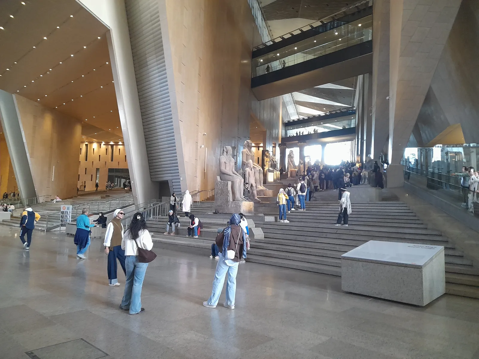 The Grand Egyptian Museum's main hall, a vast stone-clad atrium with sloping walls and a broad flight of steps rising past colossal pharaonic statues, visitors crossing the polished floor below
