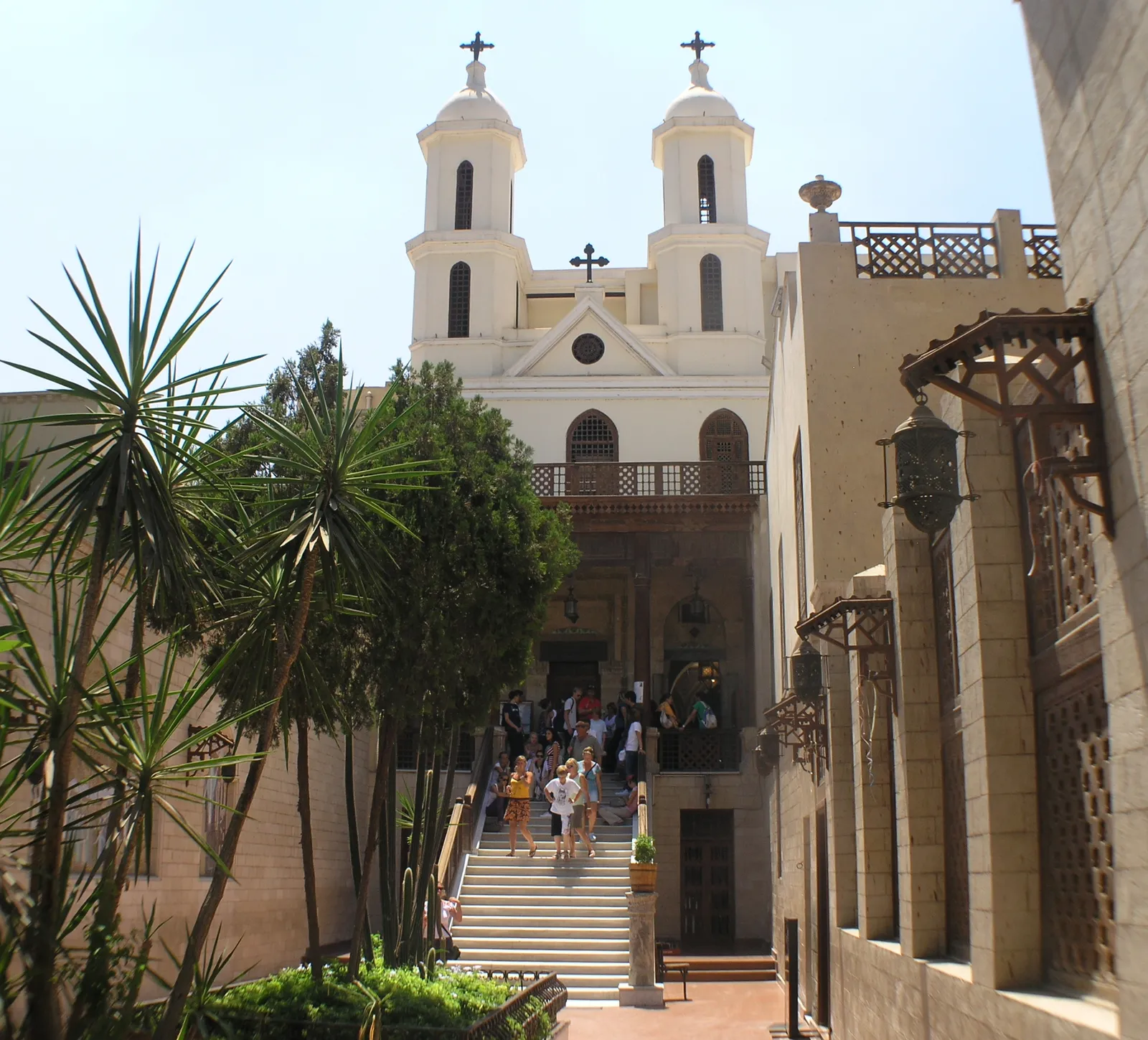 The Hanging Church in Coptic Cairo seen from its courtyard, twin white bell towers above a carved stone facade, with a flight of steps leading up to the entrance and palms in the foreground