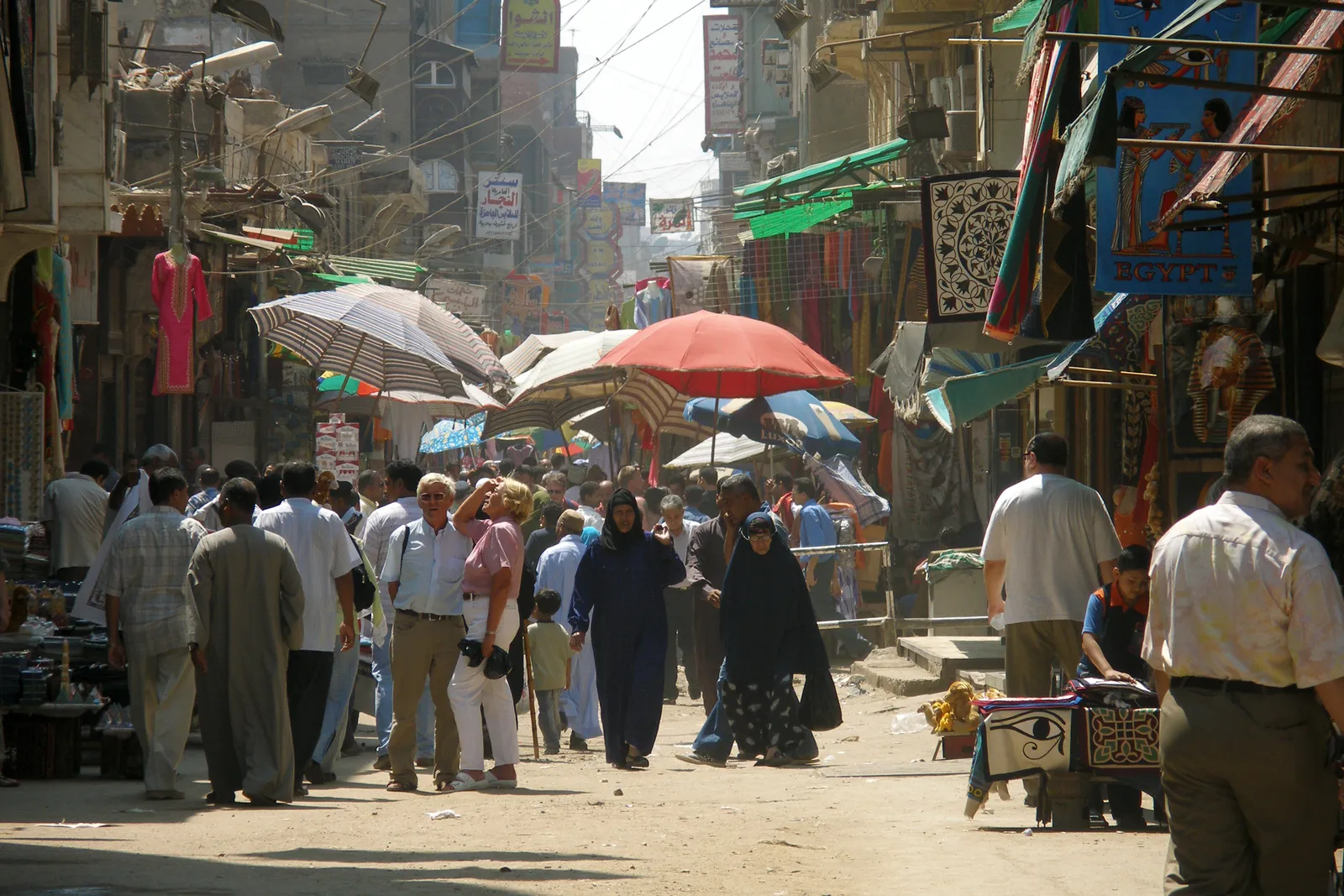 A market street in medieval Cairo packed with shoppers, parasols and canvas awnings shading the stalls on both sides and hanging fabrics strung above the shopfronts