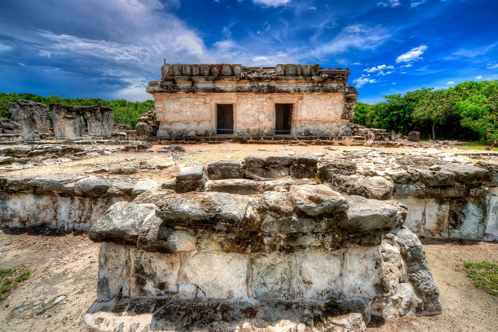 The El Rey archaeological zone in Cancún, a small stone temple with two doorways standing behind low limestone platforms and a sandy path, under a dark blue sky