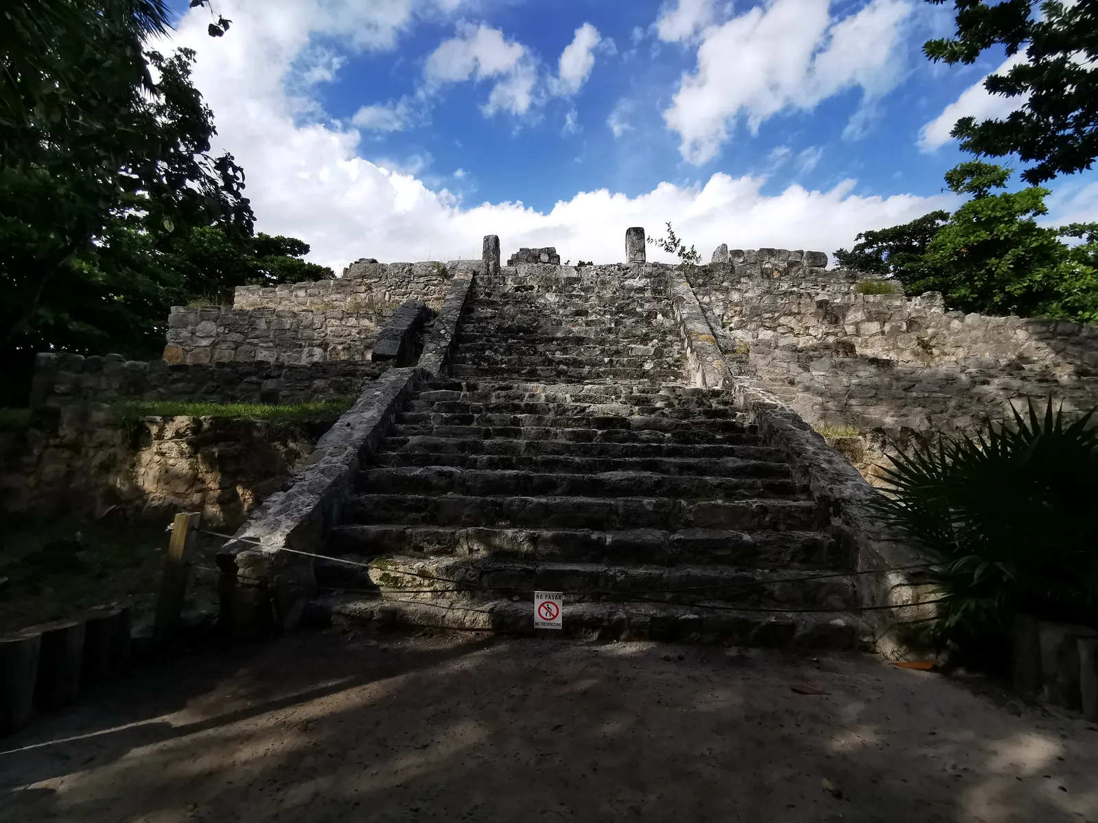 The stepped stone pyramid at the San Miguelito archaeological site in Cancún, a broad staircase running up its front, with tropical trees on either side under a cloudy blue sky