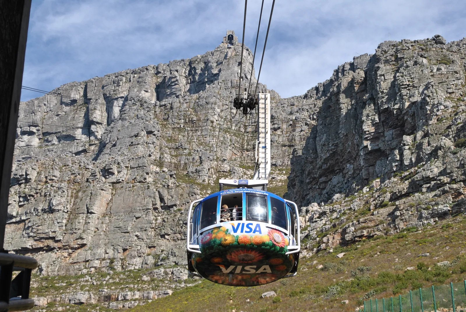 A round cable car with a band of windows hanging from its cables below the grey rock face of Table Mountain in Cape Town, the upper cable station visible on the cliff edge above