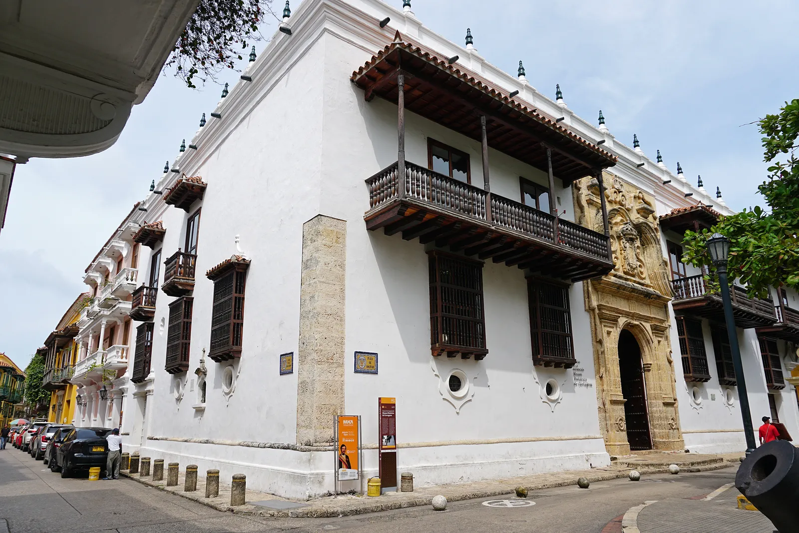 The white colonial facade of the Palacio de la Inquisicion in Cartagena, with dark wooden balconies along the upper floor and an ornate carved stone doorway at street level