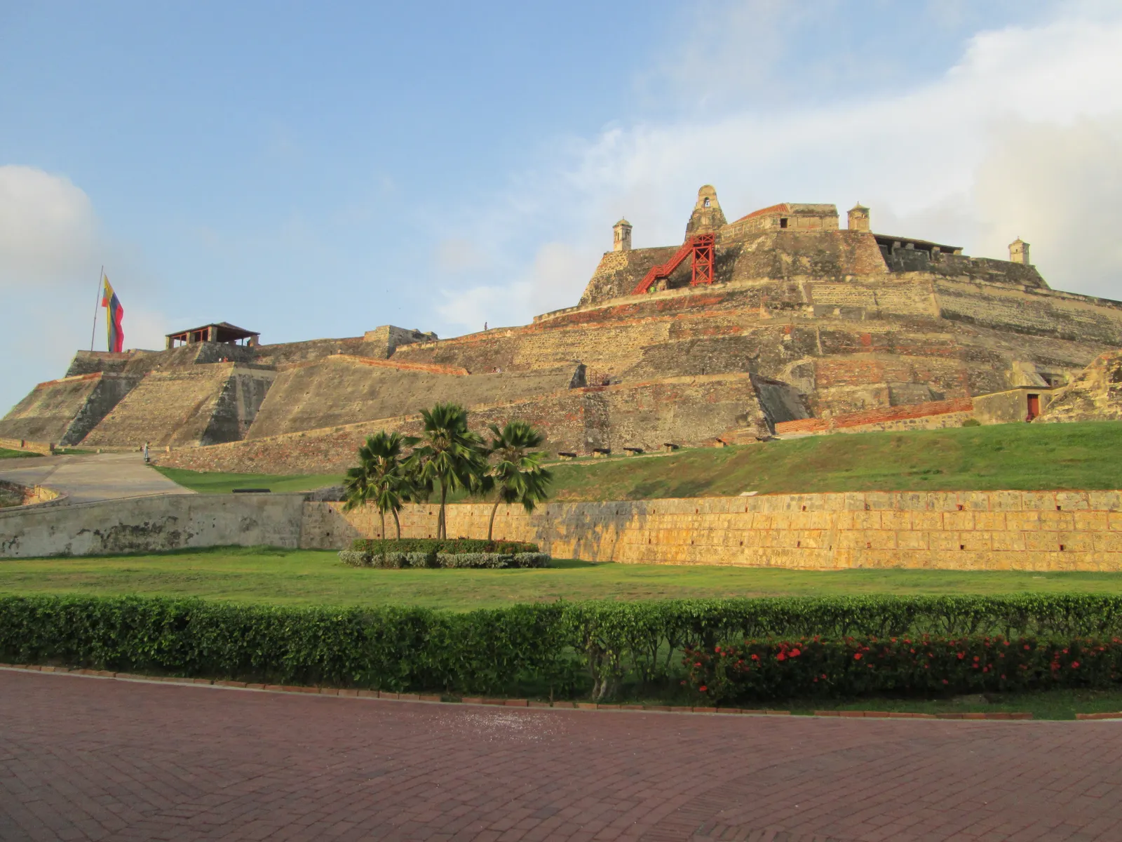 The terraced stone ramparts of Castillo San Felipe de Barajas rising on its hill in Cartagena, with a Colombian flag flying at the summit and palm trees in the foreground