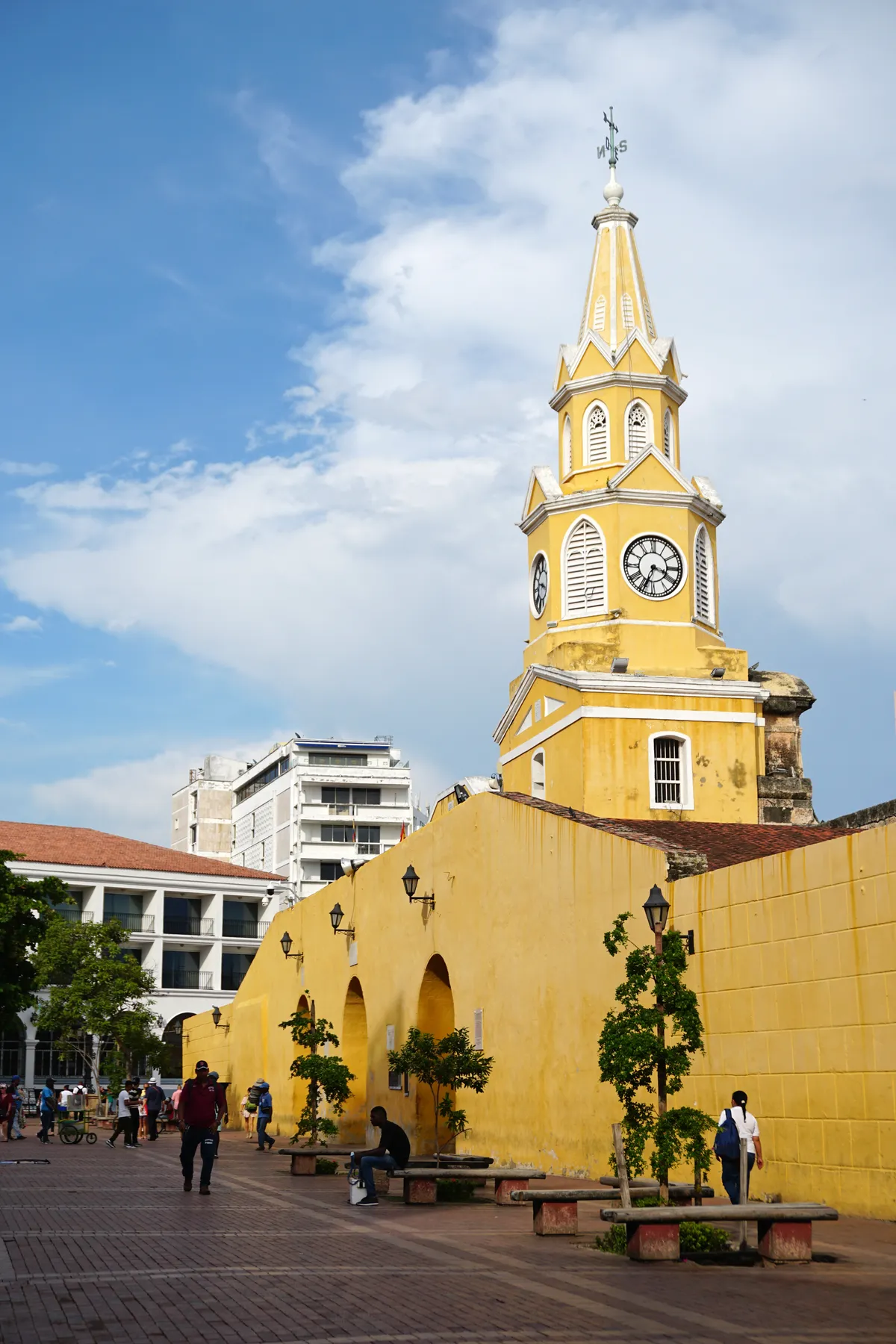The yellow Torre del Reloj clock tower gate into Cartagena's Walled City, seen from the plaza with people walking past its stone arches at dusk